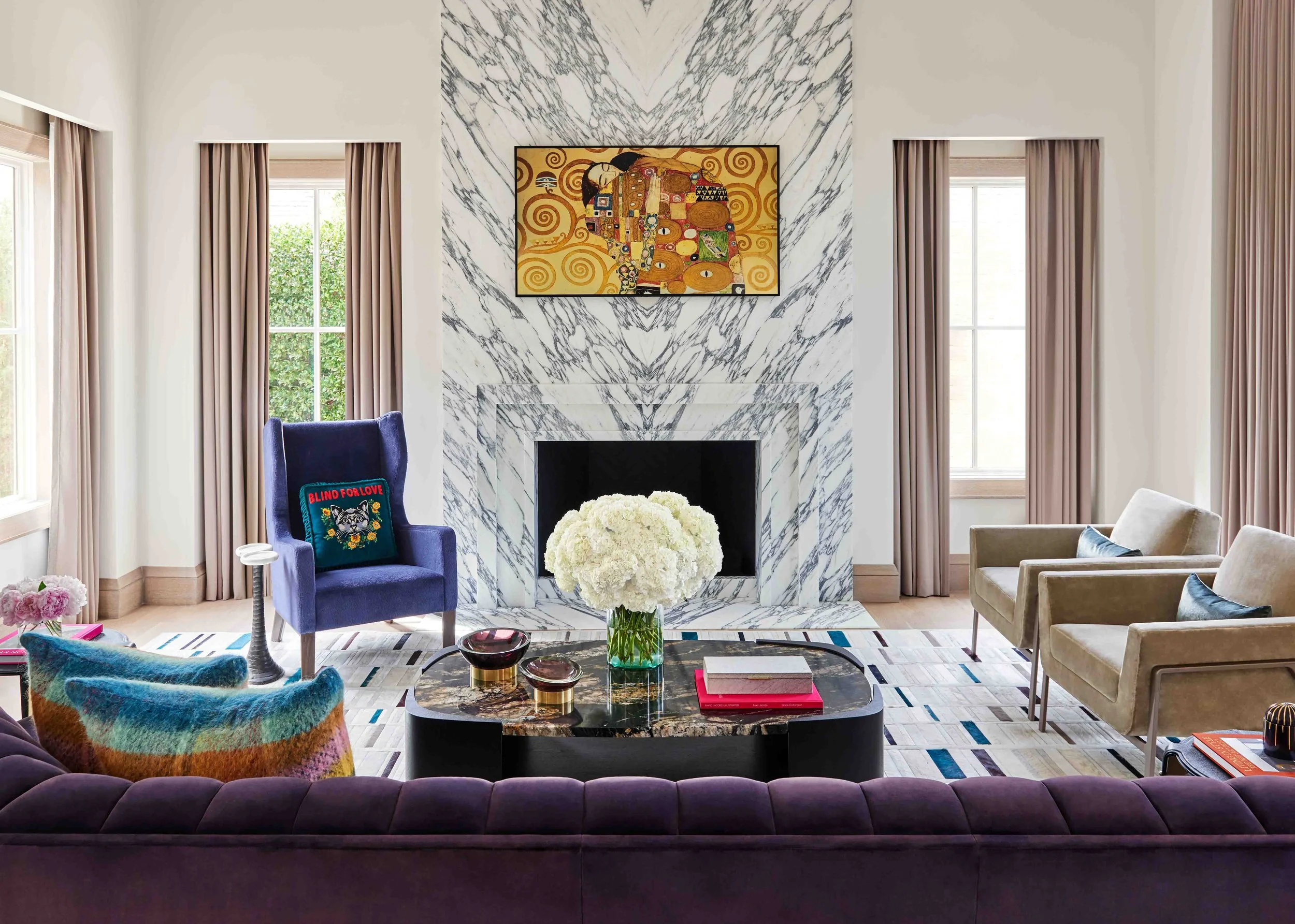 Elegant living room with a marble fireplace, abstract wall art, beige armchairs, a dark purple velvet sofa, a blue wingback chair with a Pillow that says 'Blind for Love,' colorful throw pillows, a coffee table with a bouquet of white flowers, books, and decorative bowls.