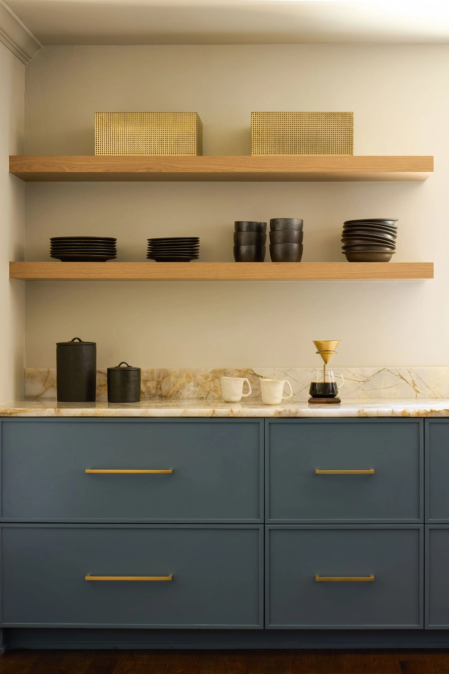 A modern kitchen cabinet with blue drawers, marble countertop, and open wooden shelves holding black and beige dishware and coffee brewing accessories.