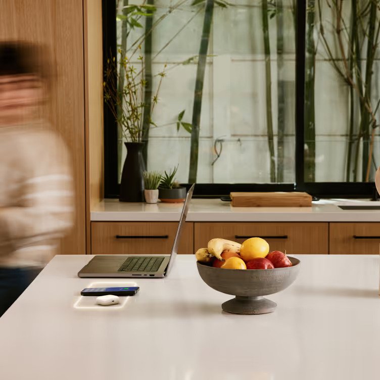 A modern kitchen with a white countertop, a laptop, a bowl of mixed fruits, and a window with bamboo plants outside.