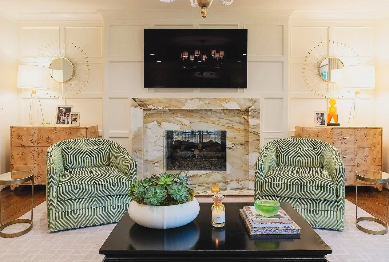 Living room with two patterned armchairs, a marble fireplace, a television above the fireplace, two wooden sideboards with lamps, framed photos, and decorative objects, a dark coffee table with a plant, candles, and a stack of books, and two round side tables.