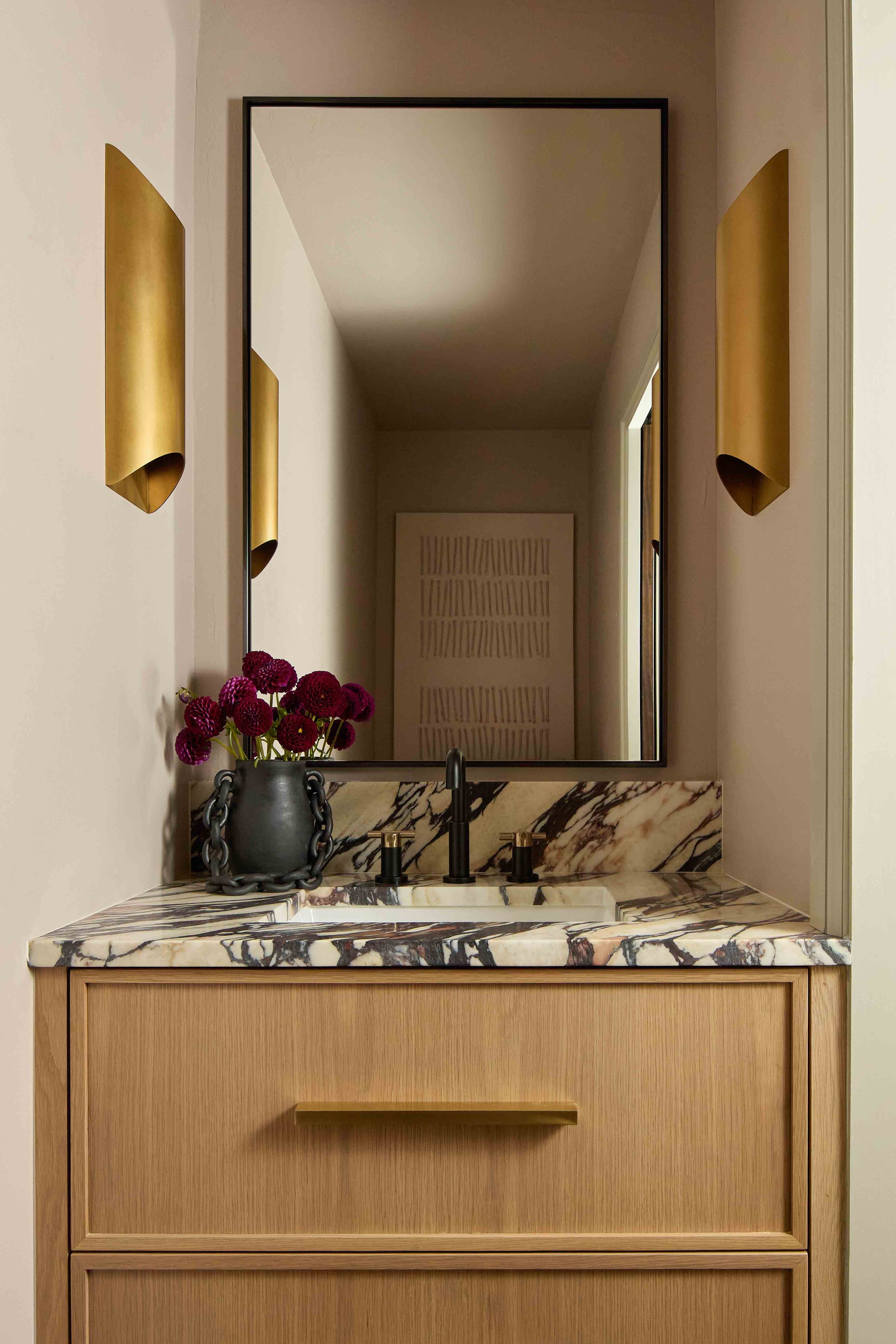 Bathroom vanity with a marble countertop, a black faucet, a dark vase with red flowers, a large mirror, and gold wall sconces.
