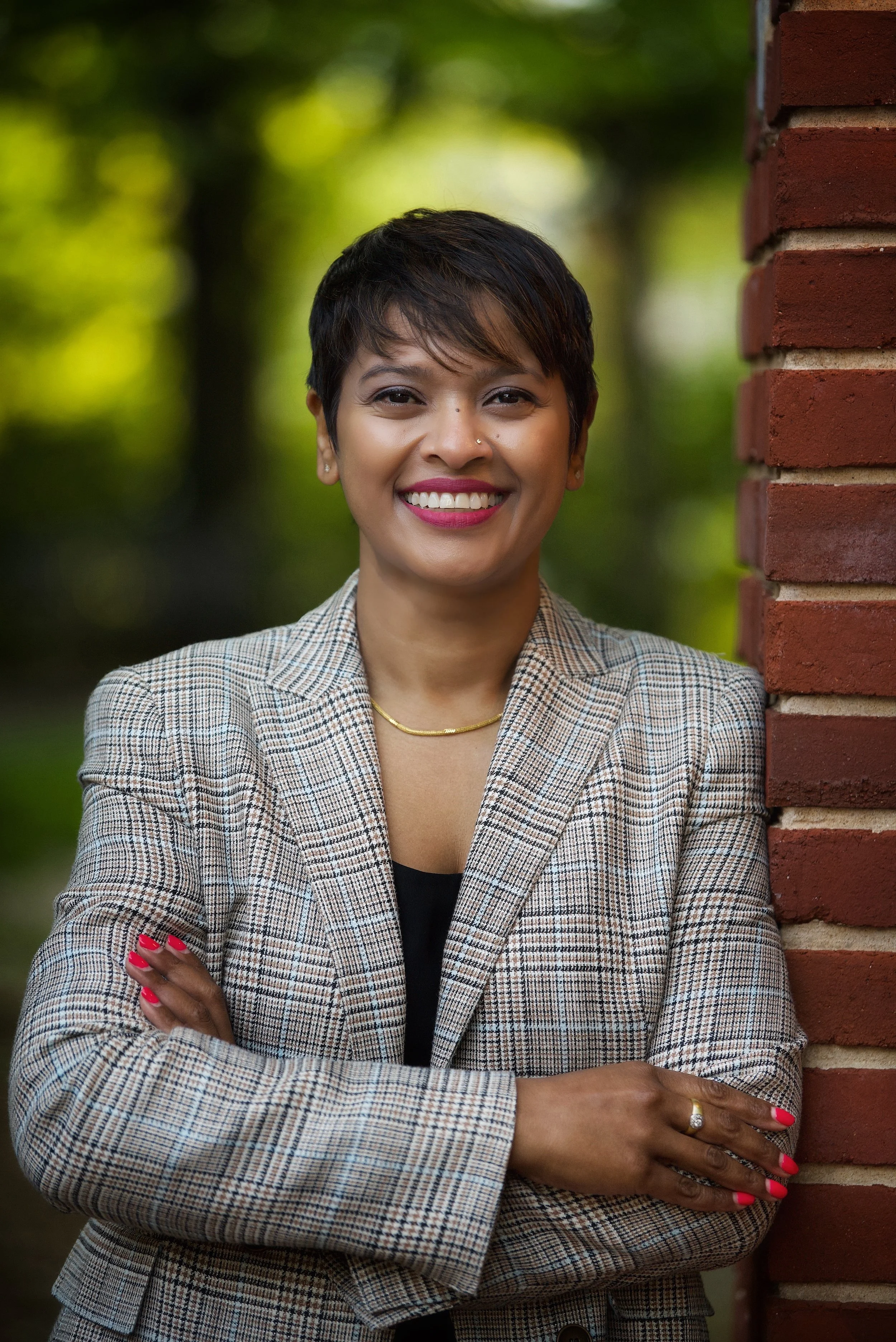 Shalini Rajoo, founder of Shalini Rajoo Advisory LLC, smiling outdoors near a brick wall with green foliage in the background.
