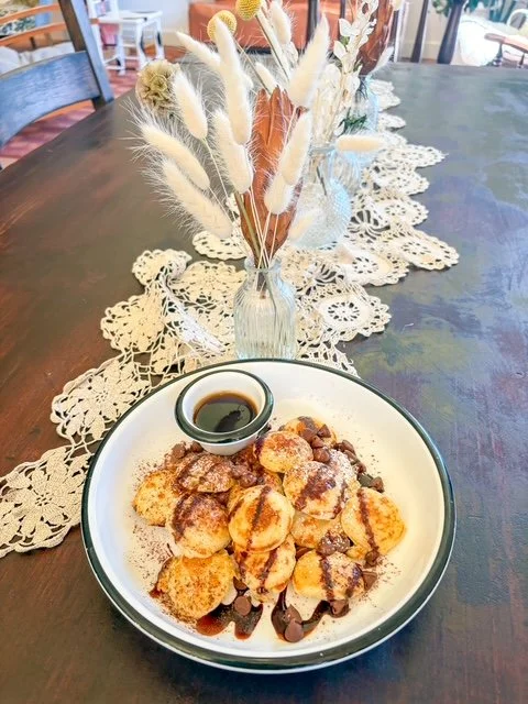A plate of dessert topped with chocolate syrup, with a small cup of syrup, on a wooden dining table with a lace table runner, and a vase of dried flowers in the background.