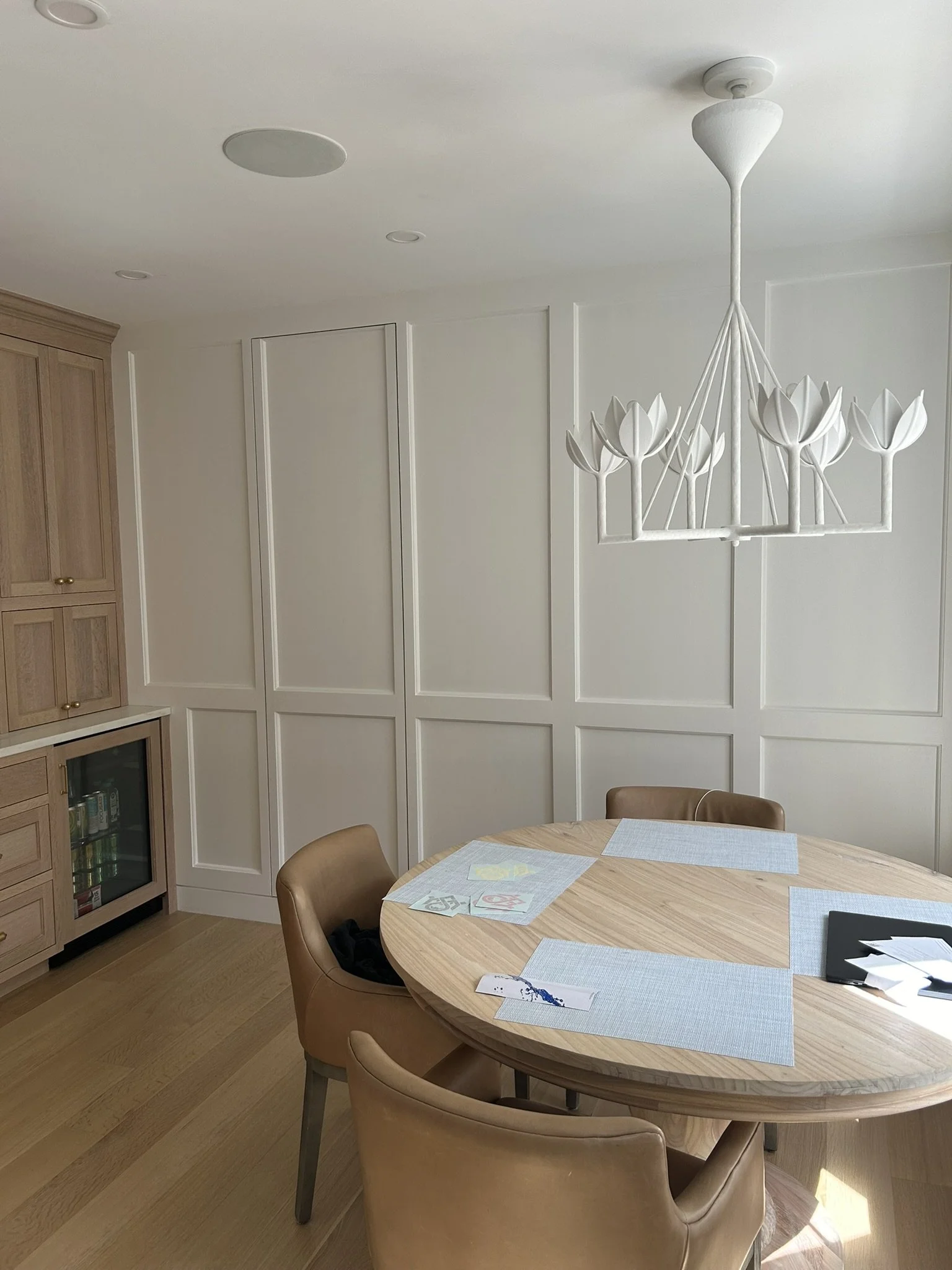 A round wooden dining table with four light brown chairs, light blue placemats, and some papers and documents. White paneled walls and a modern white chandelier with floral design hang from the ceiling in a bright, minimalistic dining room.