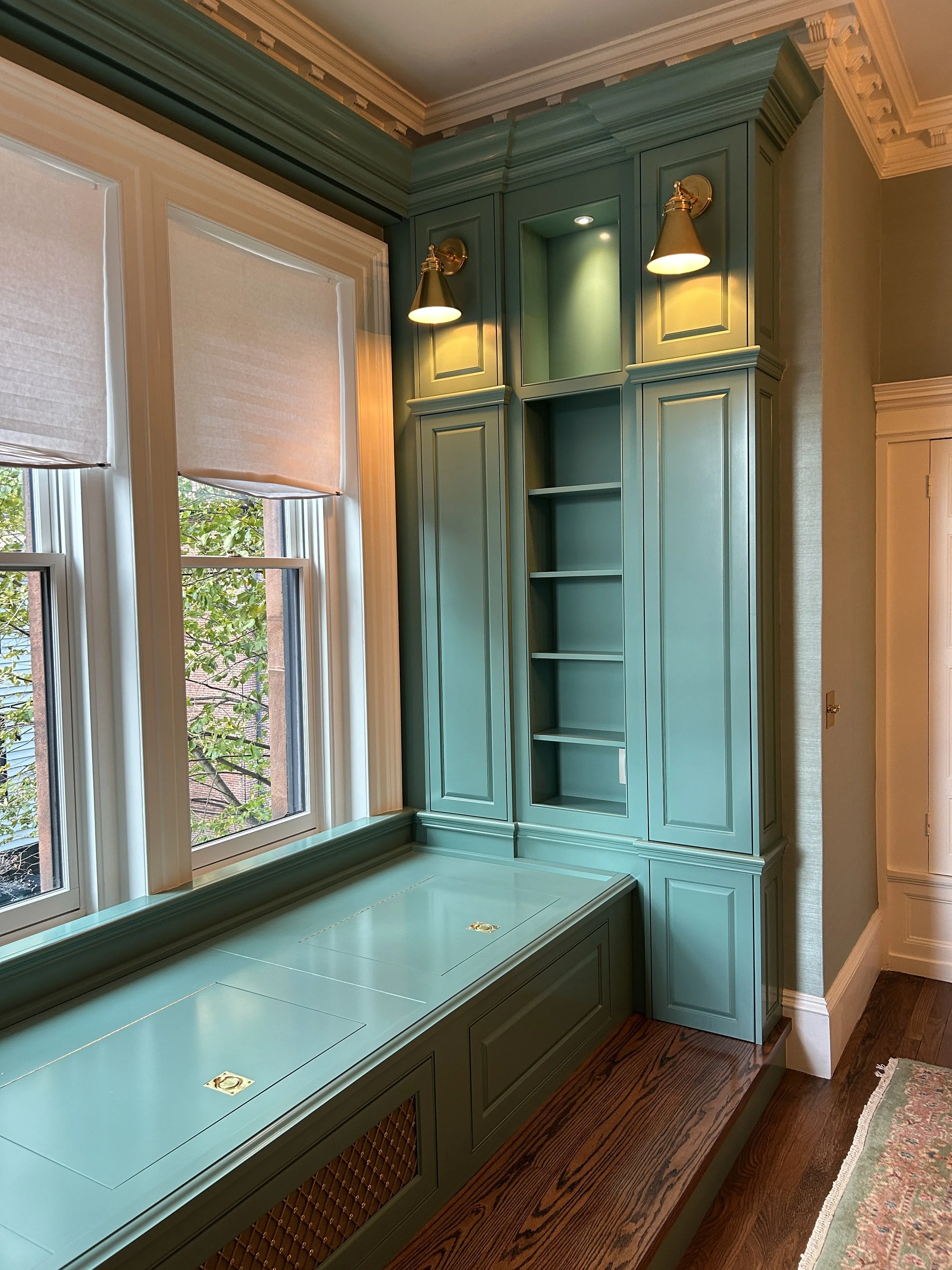 A built-in teal-colored window seat and storage cabinet with open shelves, situated next to a window with beige blinds in a room with hardwood floors and crown molding.