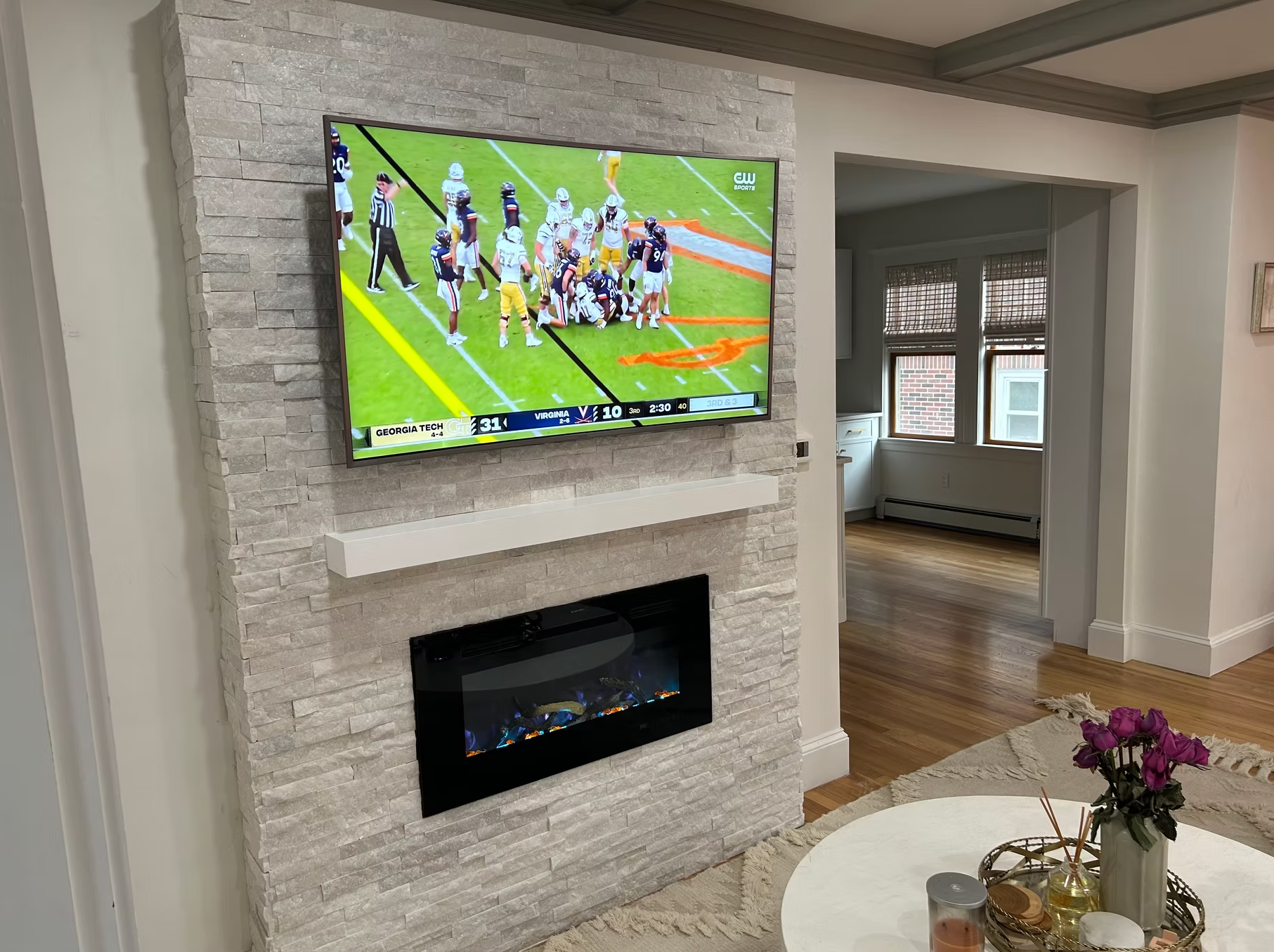 Living room with a mounted television showing a football game, a modern electric fireplace below the TV, a white floating shelf, a round coffee table with flowers, candles, and decorative objects, and a view into a bright kitchen with large windows.