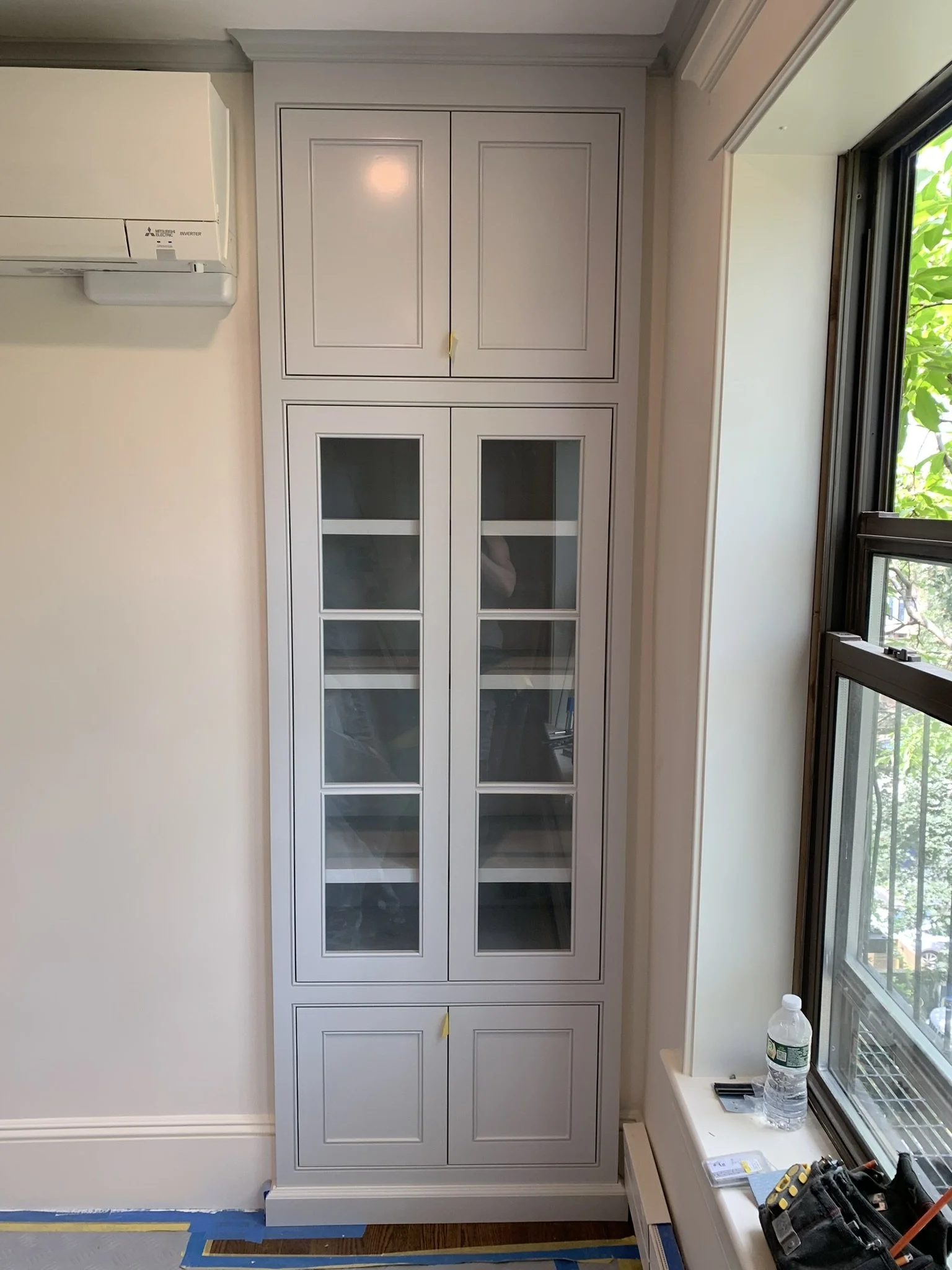 A white cabinet with glass-paneled doors on the lower section and closed doors on the upper section, placed next to a window with visible greenery outside.