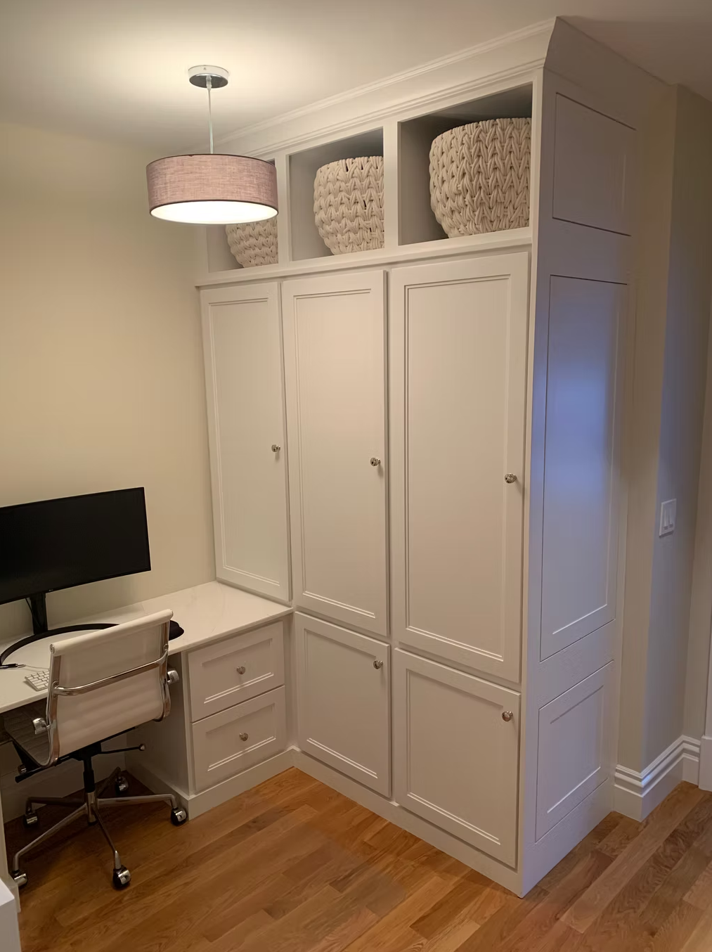 Home office corner with a built-in white cabinet, drawers, a desk with a computer monitor, and a white ergonomic chair, with woven baskets stored in open shelves above.