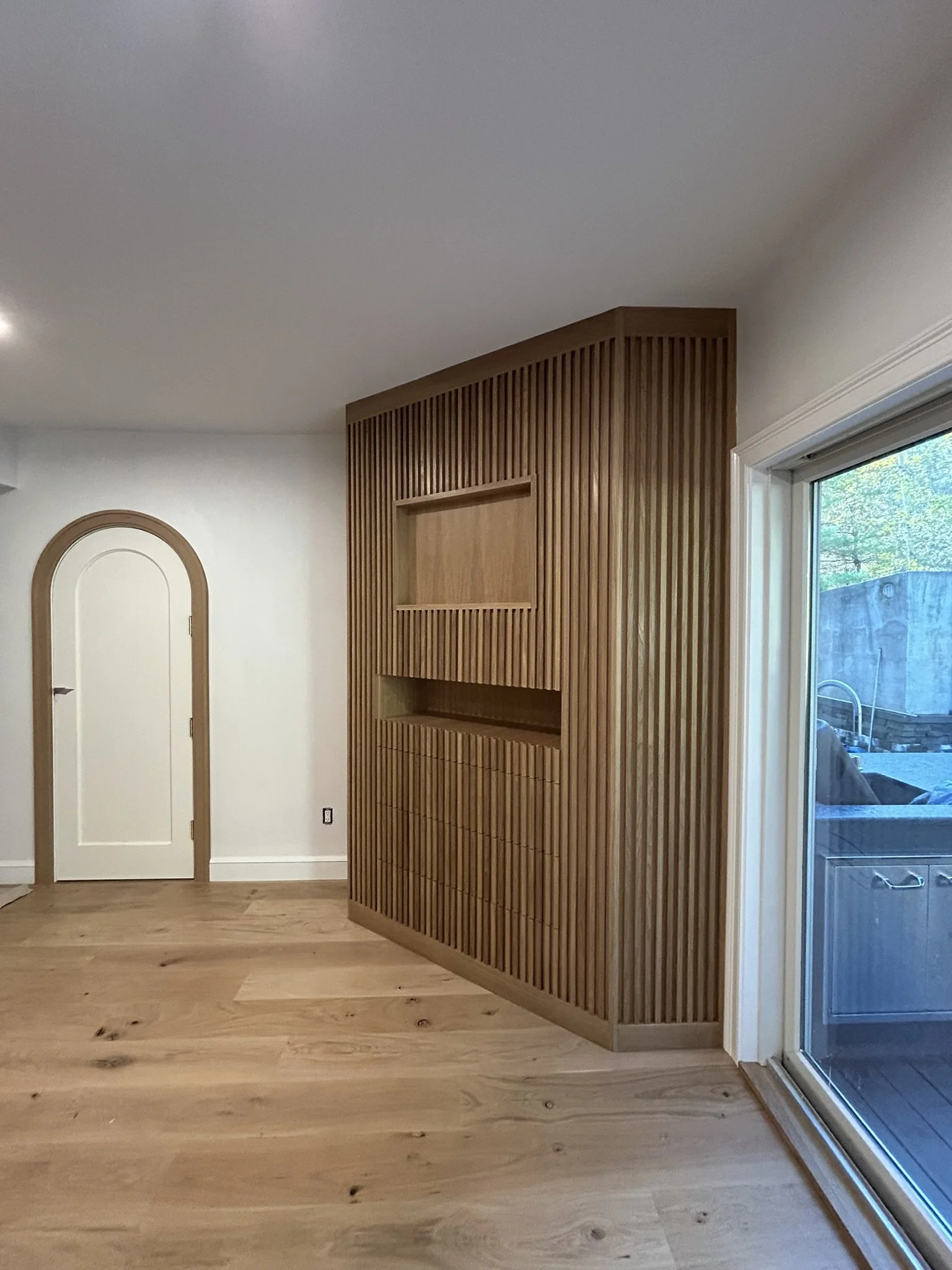 Interior of a room with light-colored hardwood flooring, a built-in wooden wall feature with vertical slats and open shelving, and a sliding glass door leading outside.