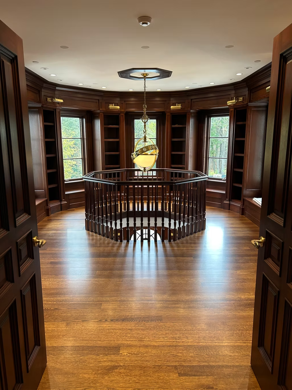 A circular wooden staircase inside a room with dark wood paneling, large windows, a hardwood floor, and a hanging light fixture in the center.