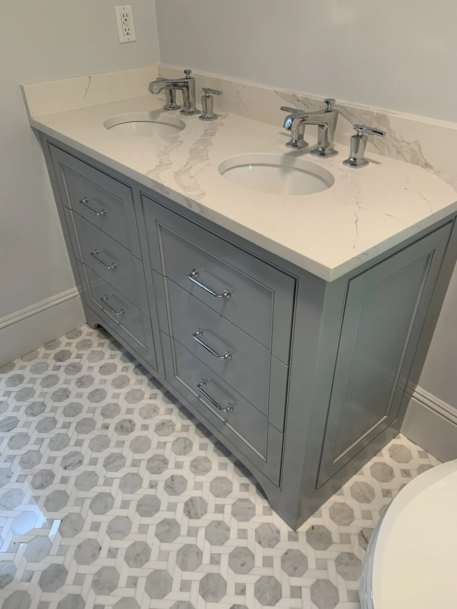 A bathroom vanity with a white marble countertop, two sinks, and chrome faucets, set against a light-colored wall with electrical outlets. The vanity has gray cabinetry with multiple drawers and silver handles. The floor features patterned white and 