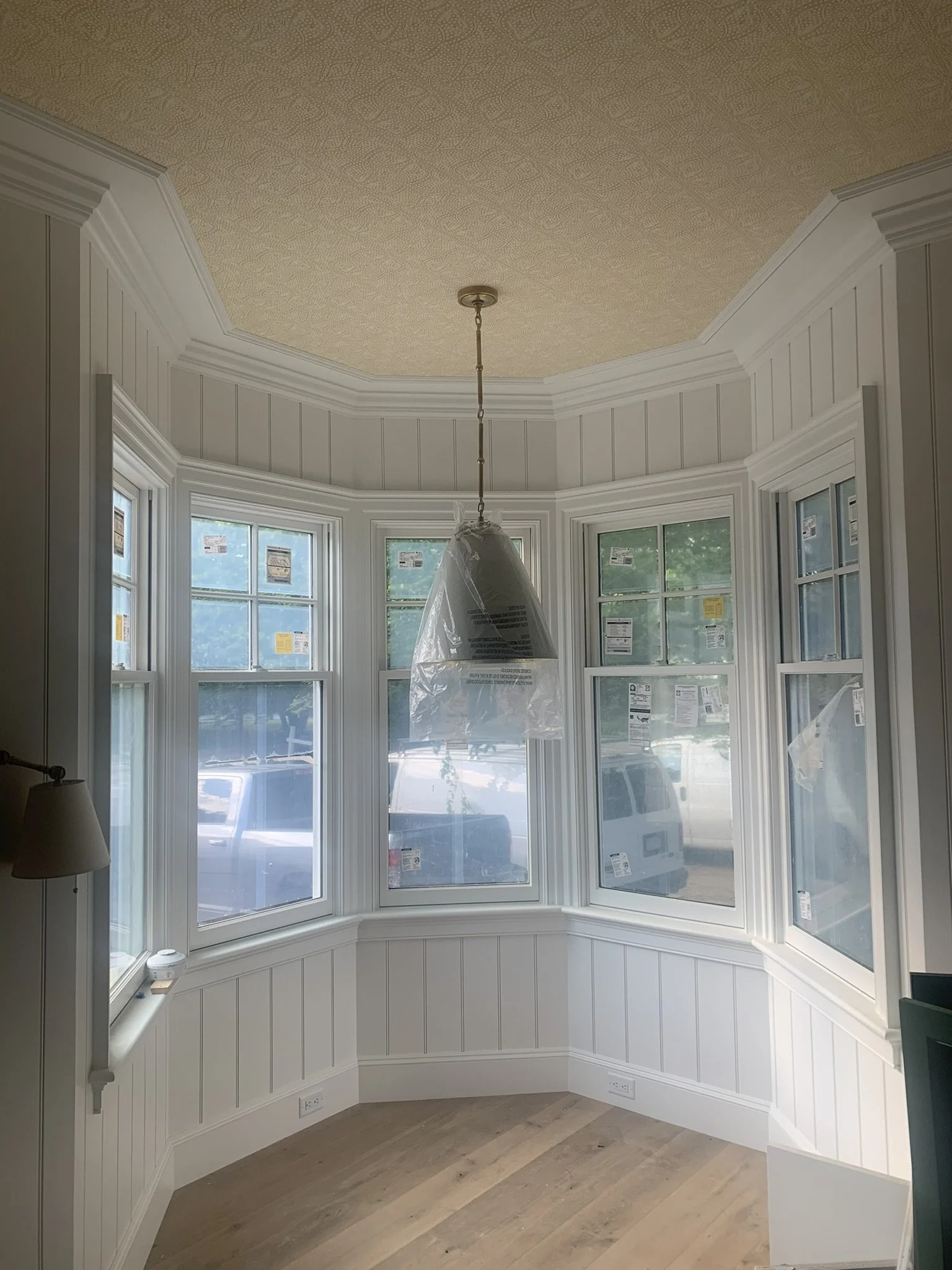 Empty room with white beadboard paneling, large bay window with multiple panes, and a hanging light fixture covered in plastic, indicating construction or renovation.
