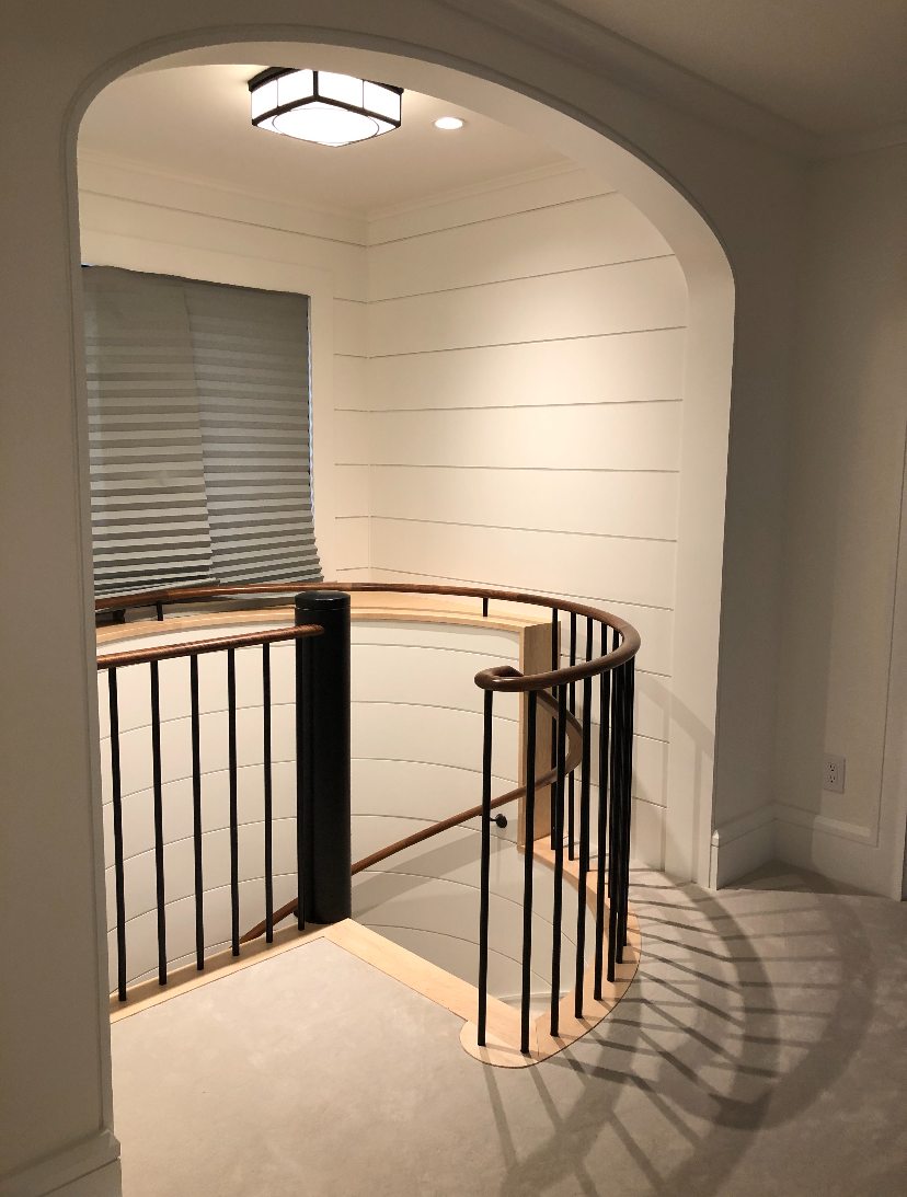 Interior view of a staircase with a curved black metal railing, light wood handrail, white shiplap walls, a window with horizontal blinds, and a ceiling light fixture.
