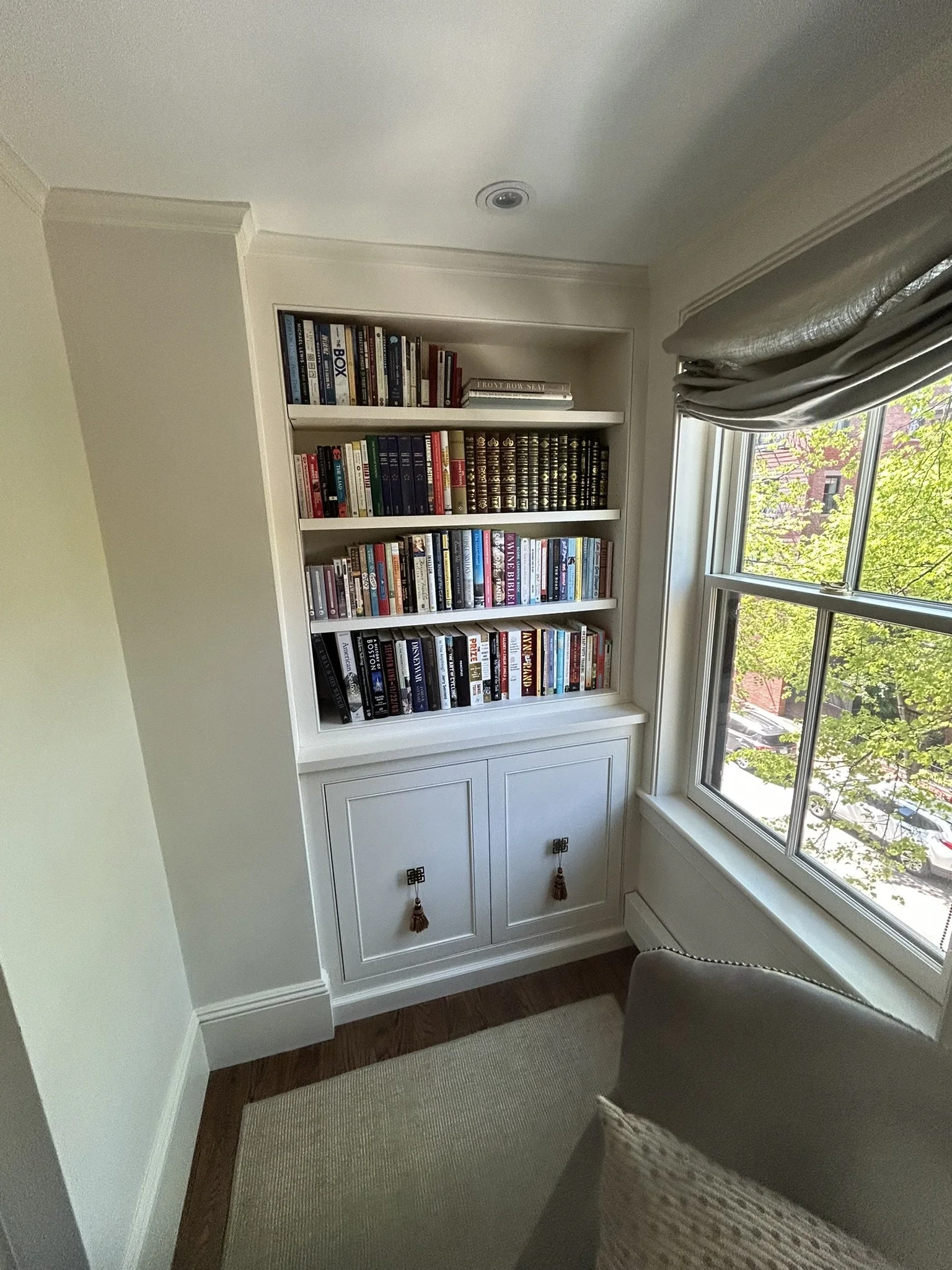 A white built-in bookshelf filled with books, located next to a window with a silver curtain, in a room with cream walls and wooden flooring.