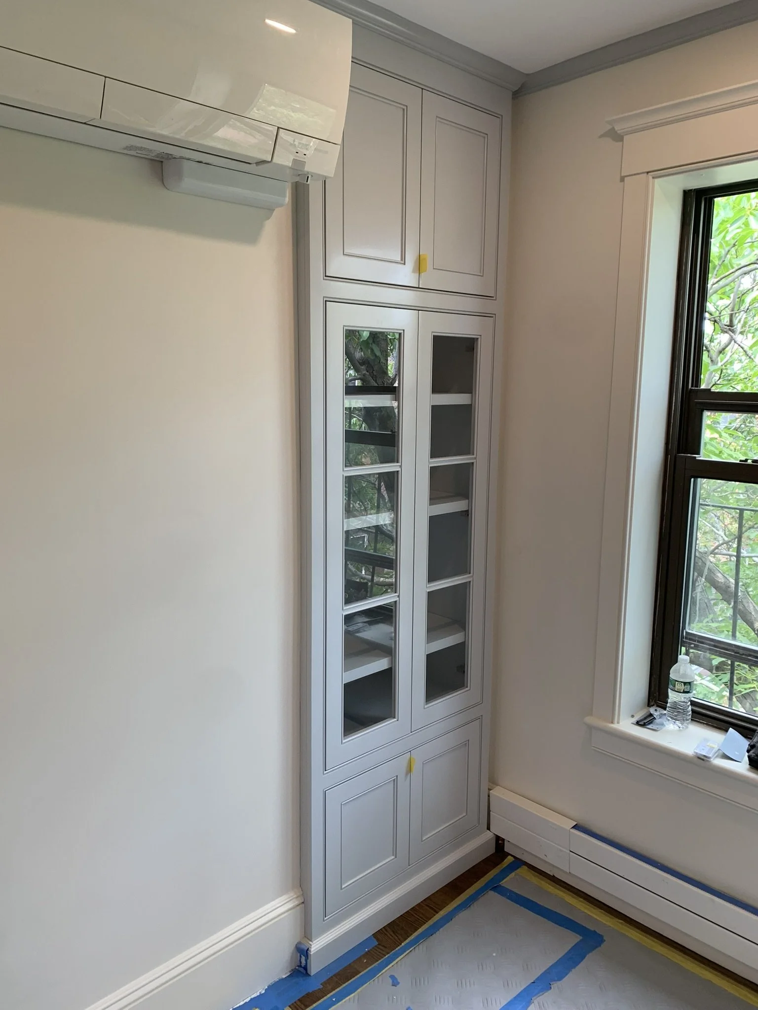 White built-in cabinet with glass-paneled doors in a room with a window and heater, some tape on the floor and cabinet, and a water bottle on the window sill.
