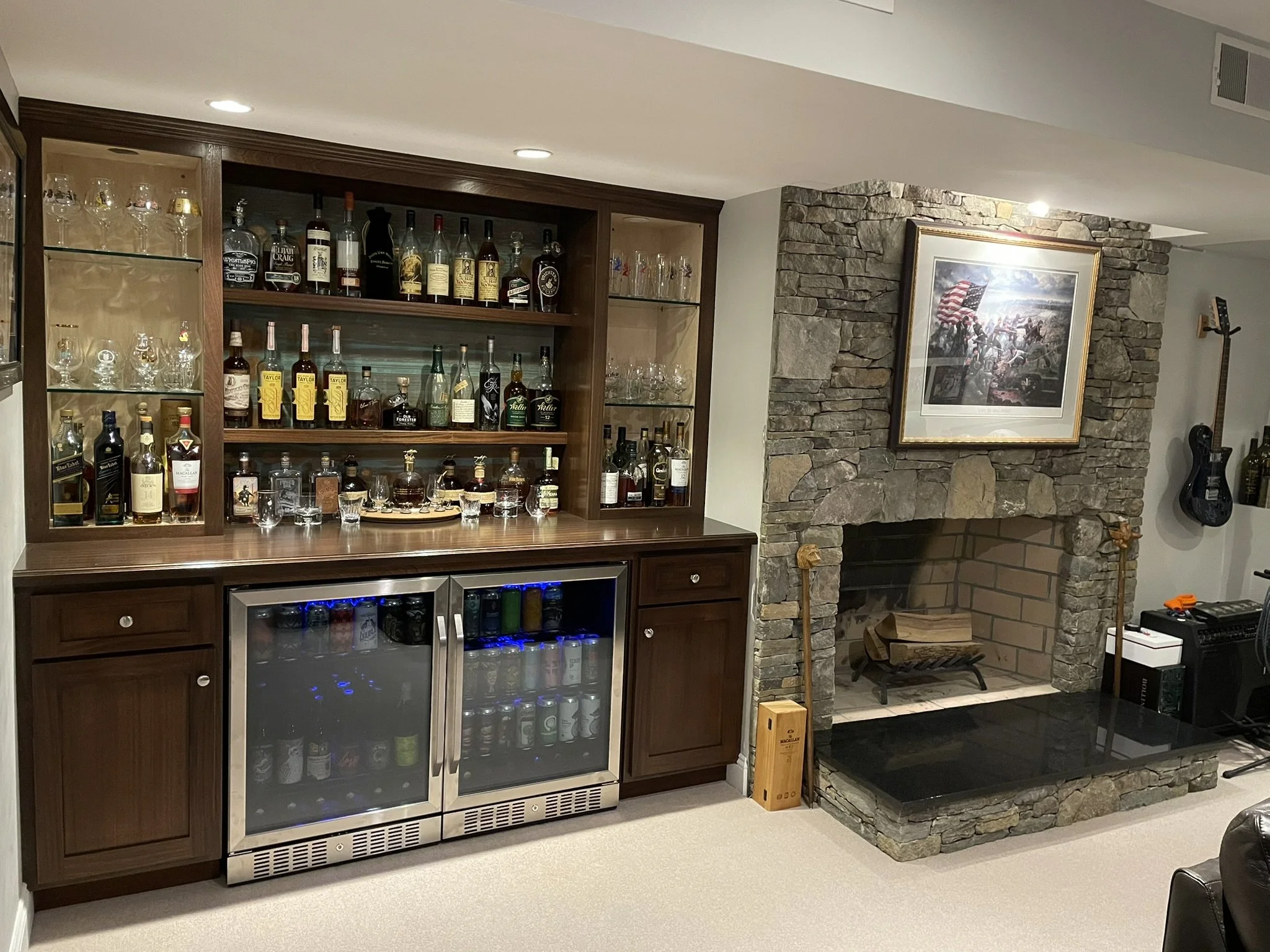 Home bar with shelves of various liquor bottles, glassware, and a beverage cooler, next to a stone fireplace with a framed painting and a wood fire grate.