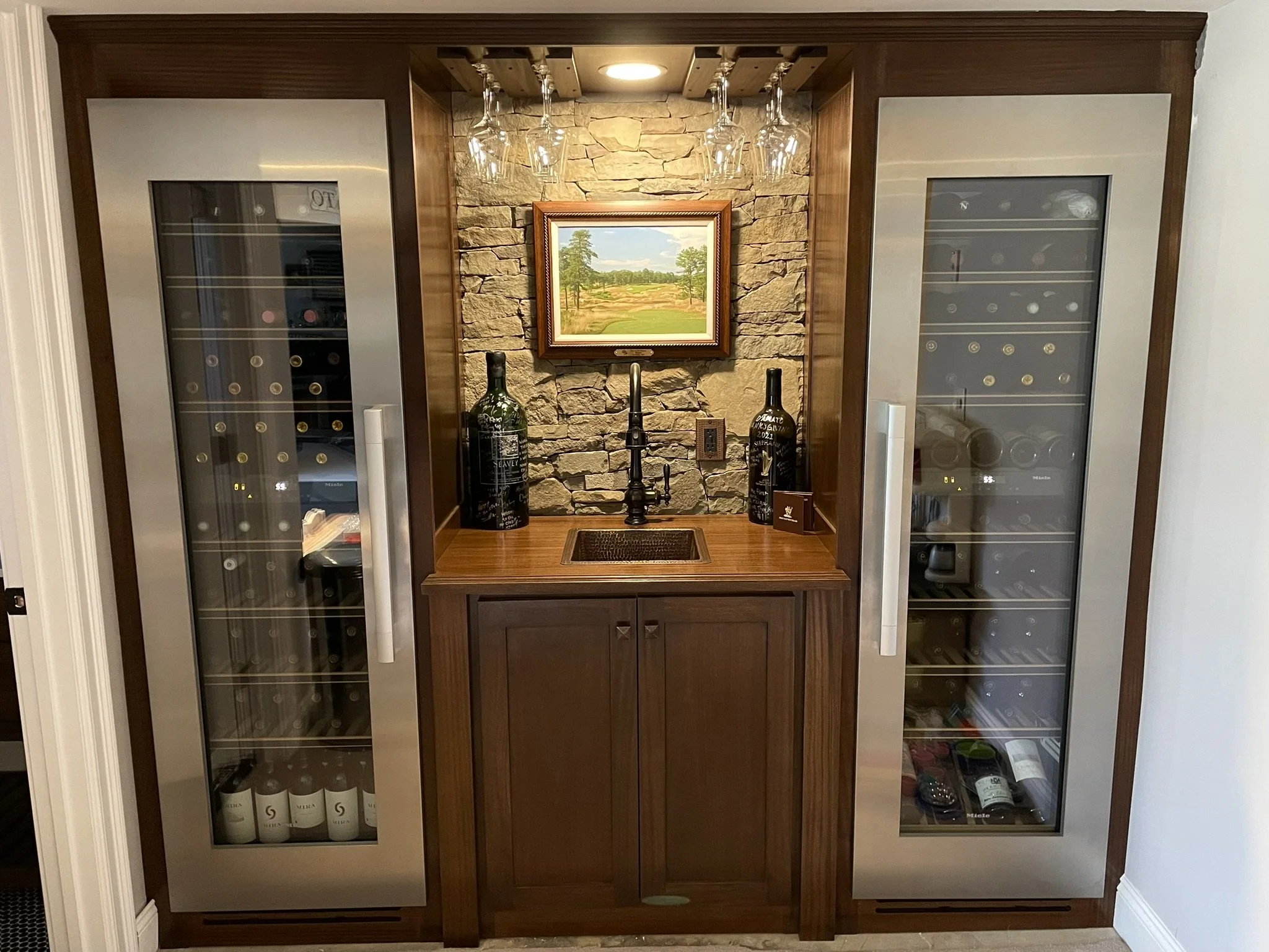 A wine bar with a stone accent wall, two wine refrigerators flanking a small wooden sink area with a framed landscape painting and hanging wine glasses.
