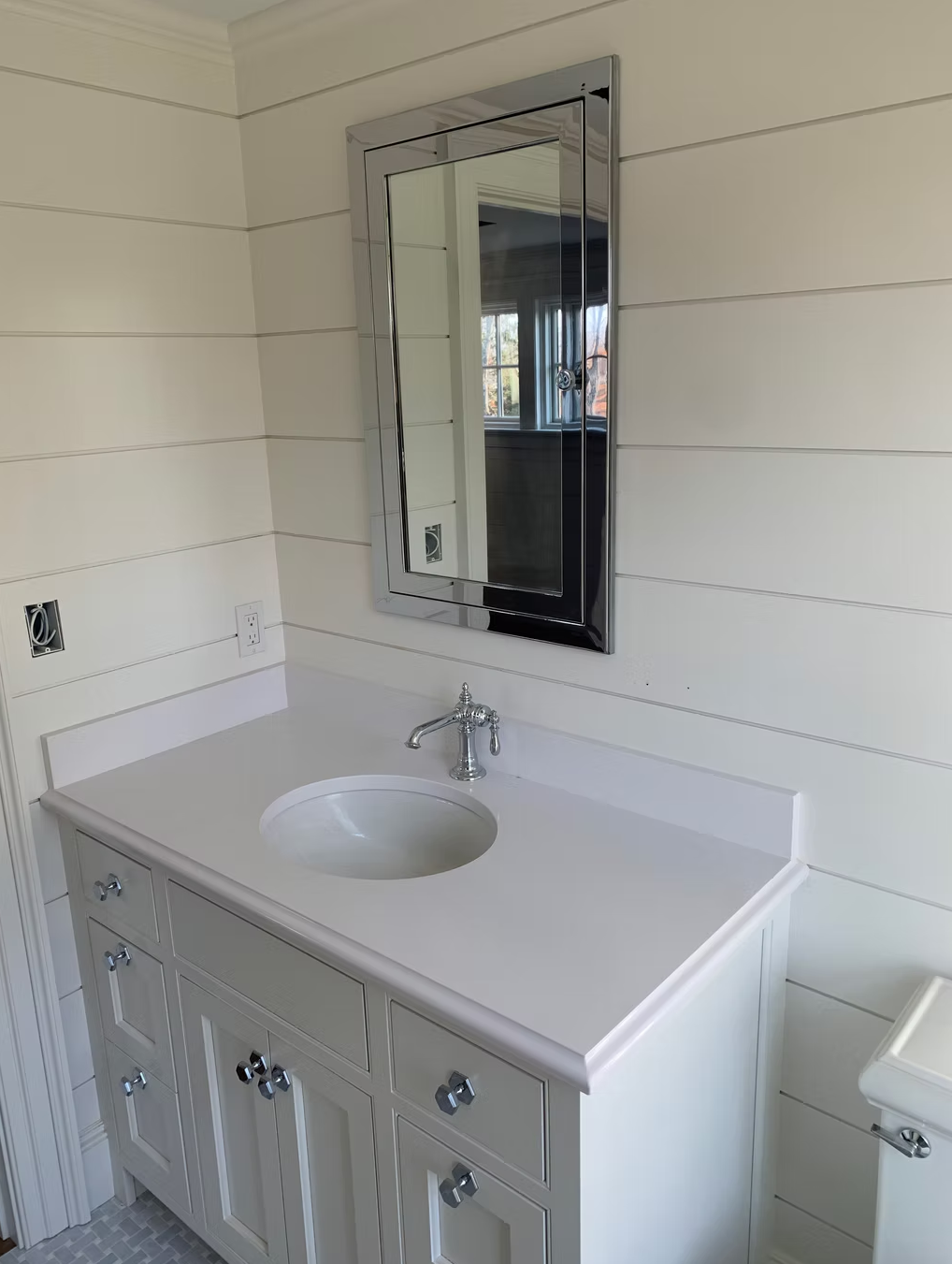 Bathroom vanity with a white countertop, single sink, and cabinet doors with crystal knobs, above a tiled floor. A rectangular wall mirror with a silver frame hangs above the vanity. The wall behind is painted with horizontal shiplap paneling.