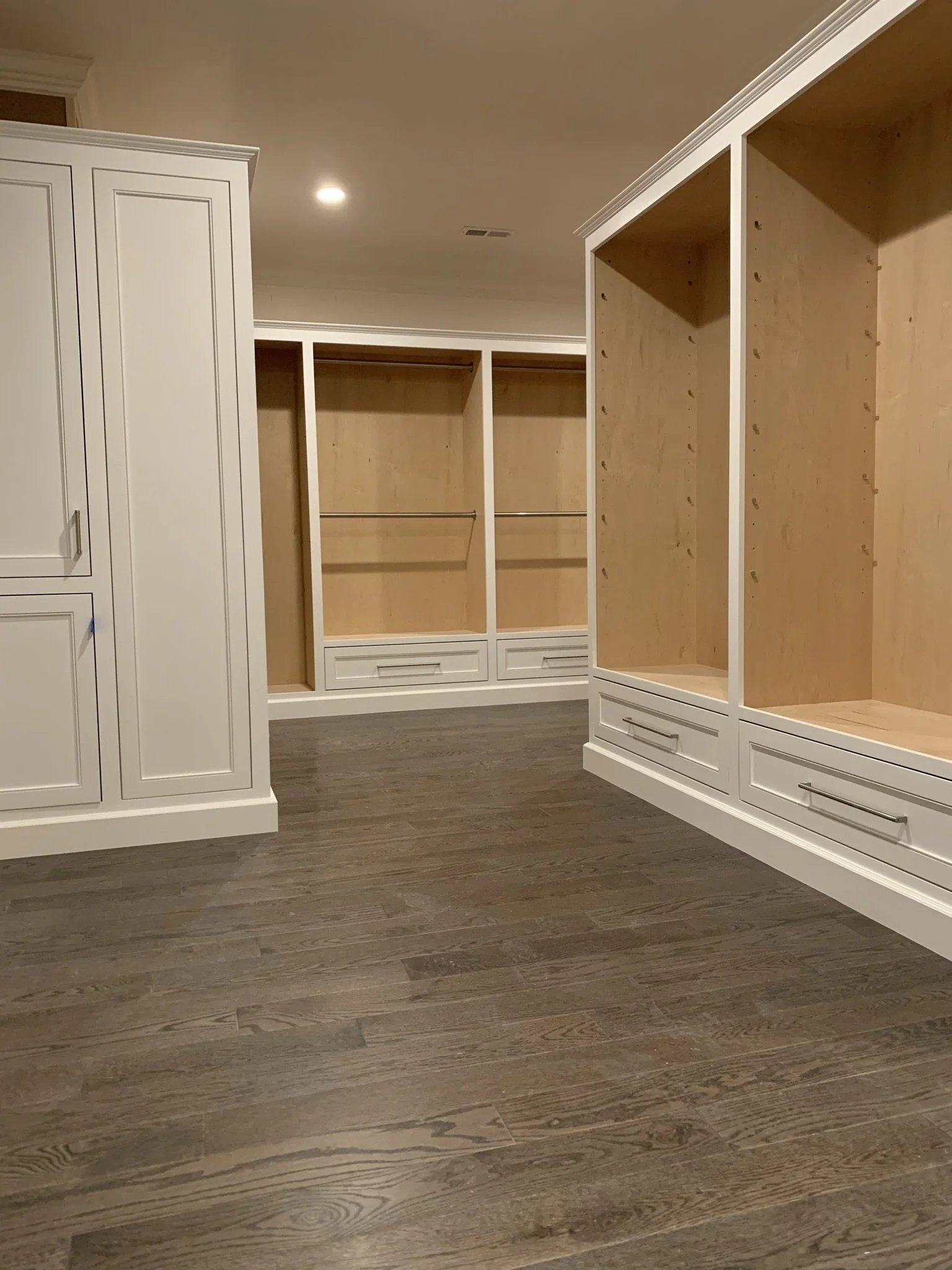 Empty walk-in closet with white built-in cabinets and drawers, wooden flooring, and unfinished shelving units.