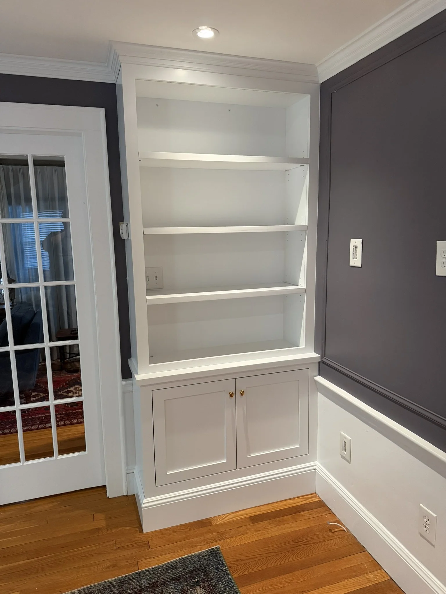 Empty white built-in bookshelf with three shelves and a cabinet with two doors at the bottom, located in a room with hardwood floors and gray walls.
