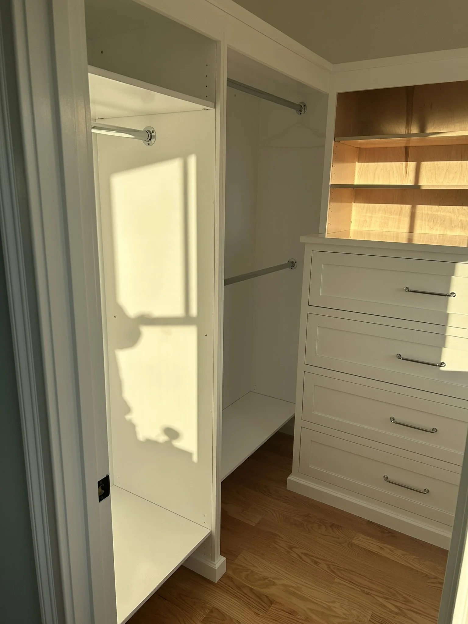 Empty walk-in closet with white shelving, drawers, and hanging rods, illuminated by sunlight.