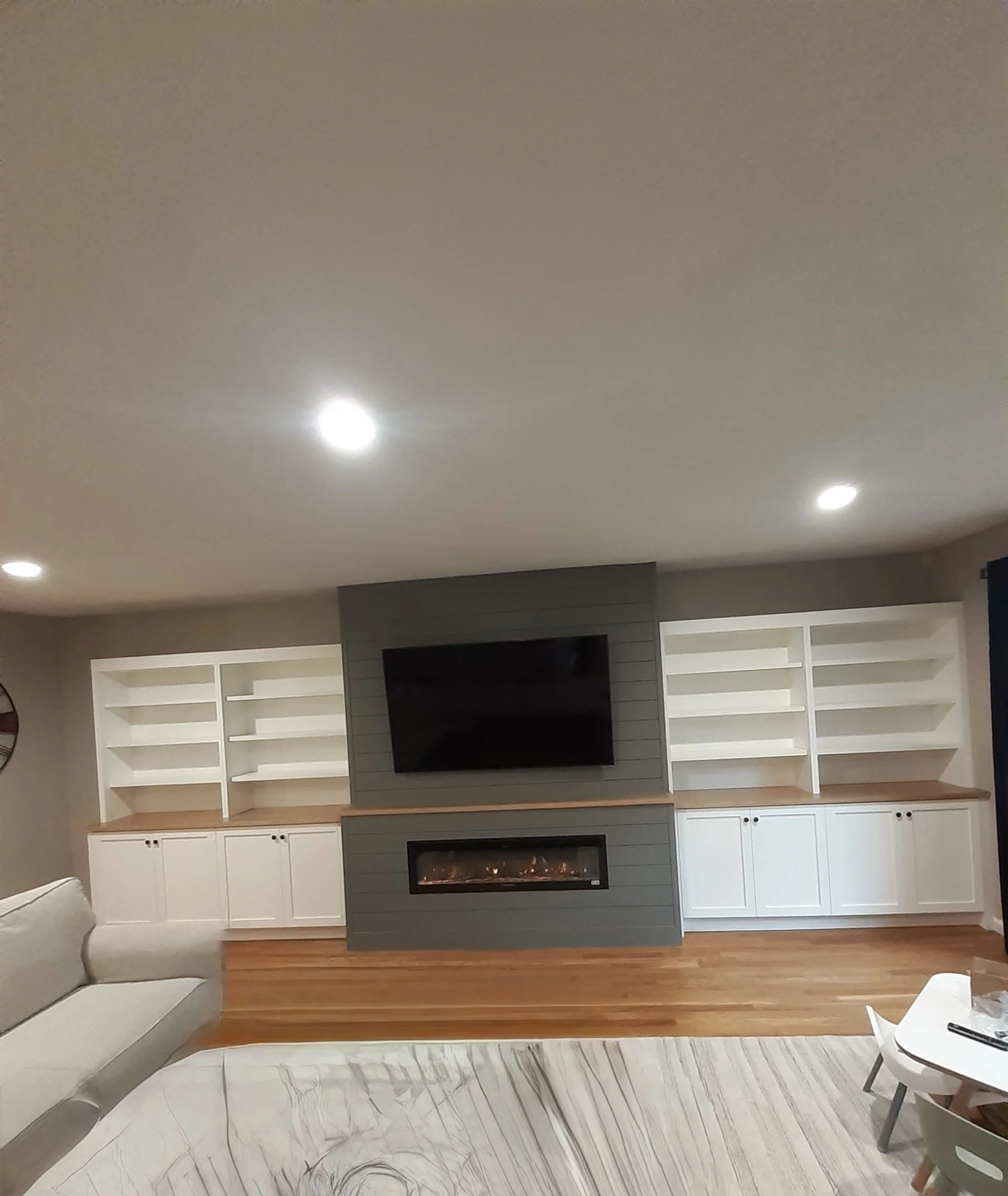 Living room with built-in white shelves, a gray center wall with a mounted television and electric fireplace, hardwood flooring, part of a white sofa, and a dining table with chairs.