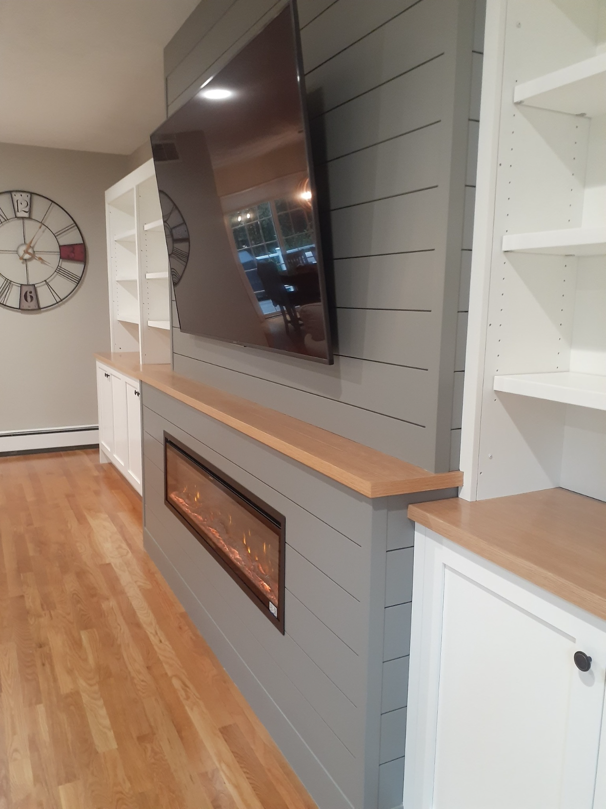 Living room feature wall with a wall-mounted TV above a modern electric fireplace, white built-in shelves, and a wooden cabinet on a gray shiplap accent wall, with hardwood floors.