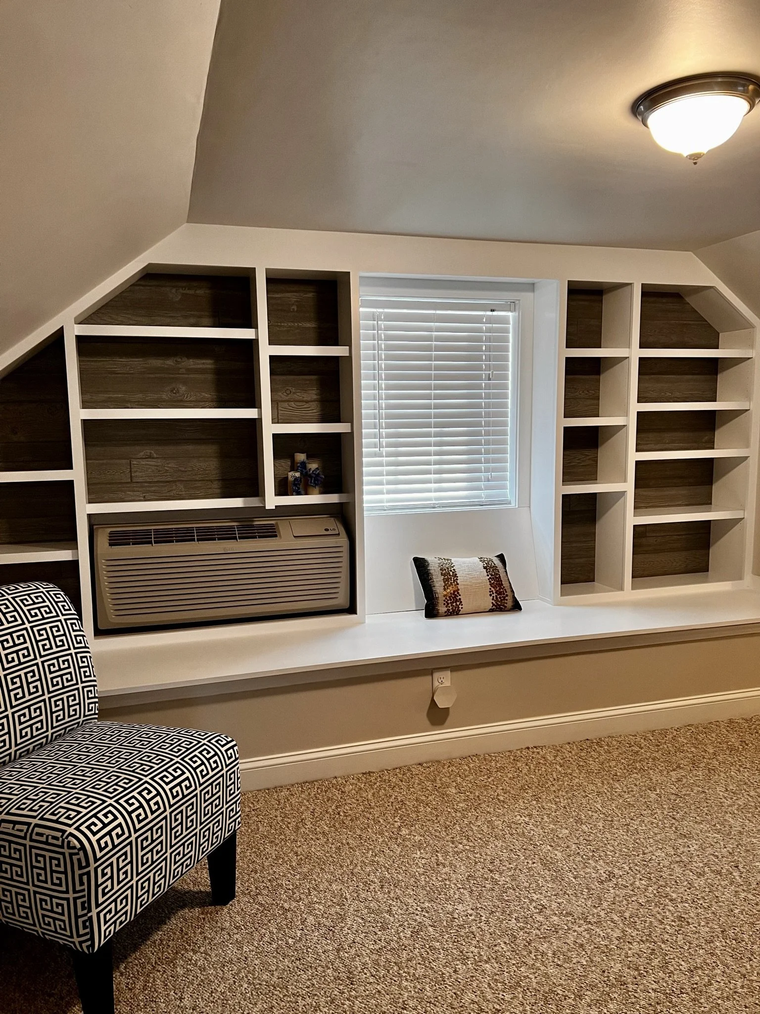 A built-in bookshelf with a white frame and wood backing, located under a sloped ceiling with a window with blinds. There is a cushioned pillow on the window seat, a patterned chair in the foreground, and a ceiling light.
