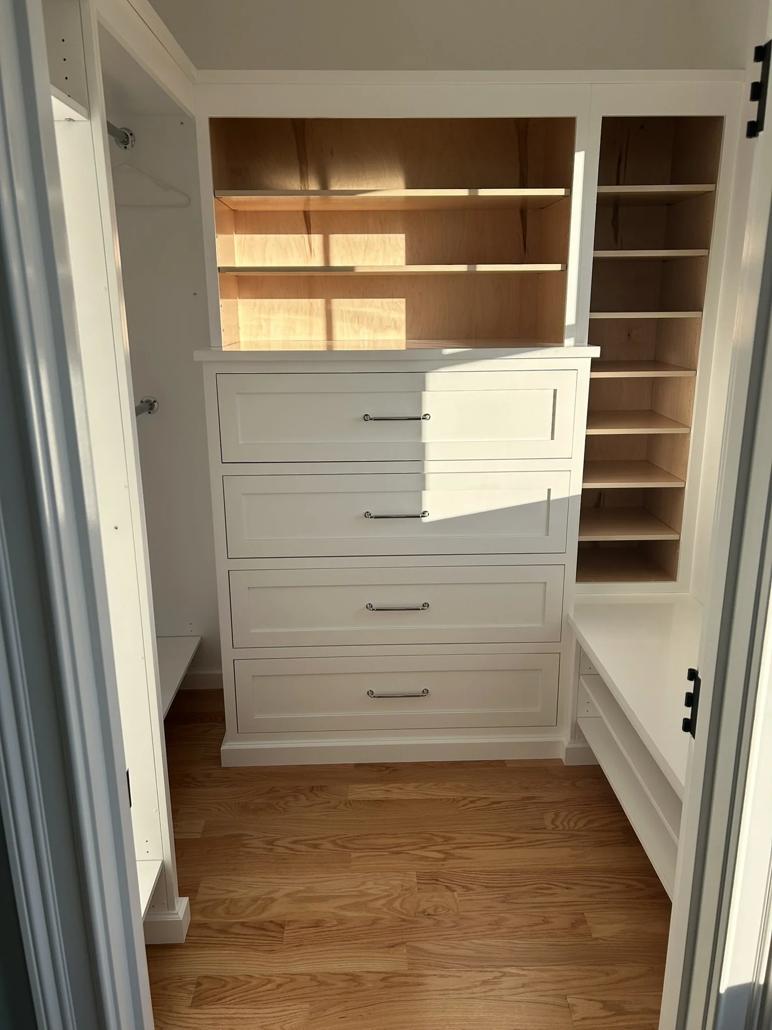 Empty custom closet with white drawers, open shelves, and light wood accents, illuminated by sunlight.