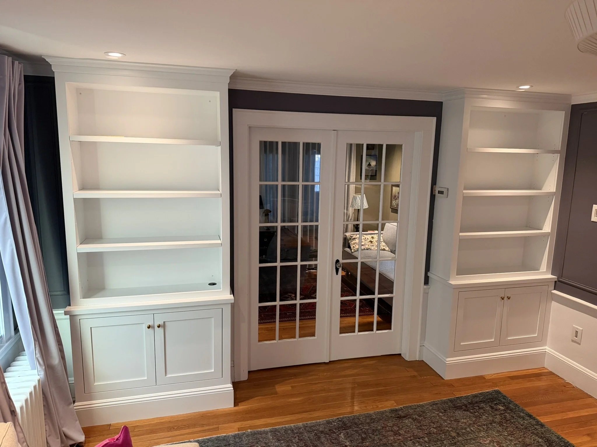 Empty white bookshelves on either side of a glass-paned double door, revealing a room with a bed and nightstand in the background. Hardwood floors and painted walls are visible.