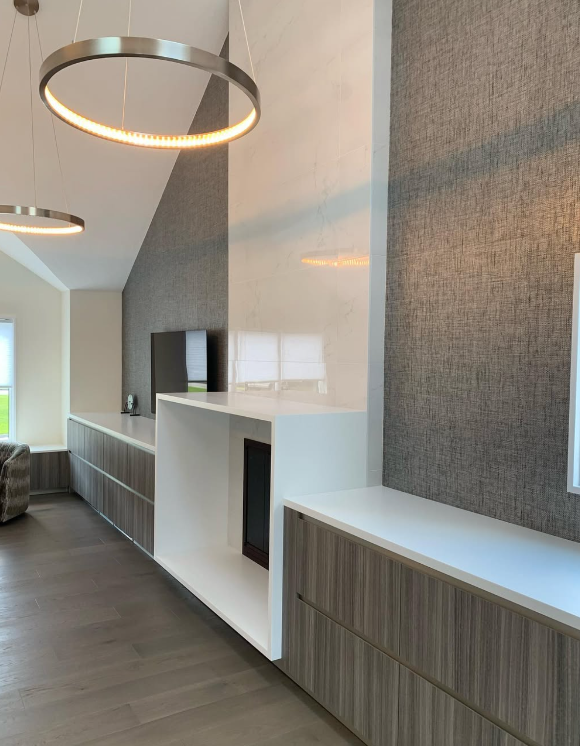 Modern living room with a wall-mounted television, textured grey walls, and wooden cabinetry beneath large windows.