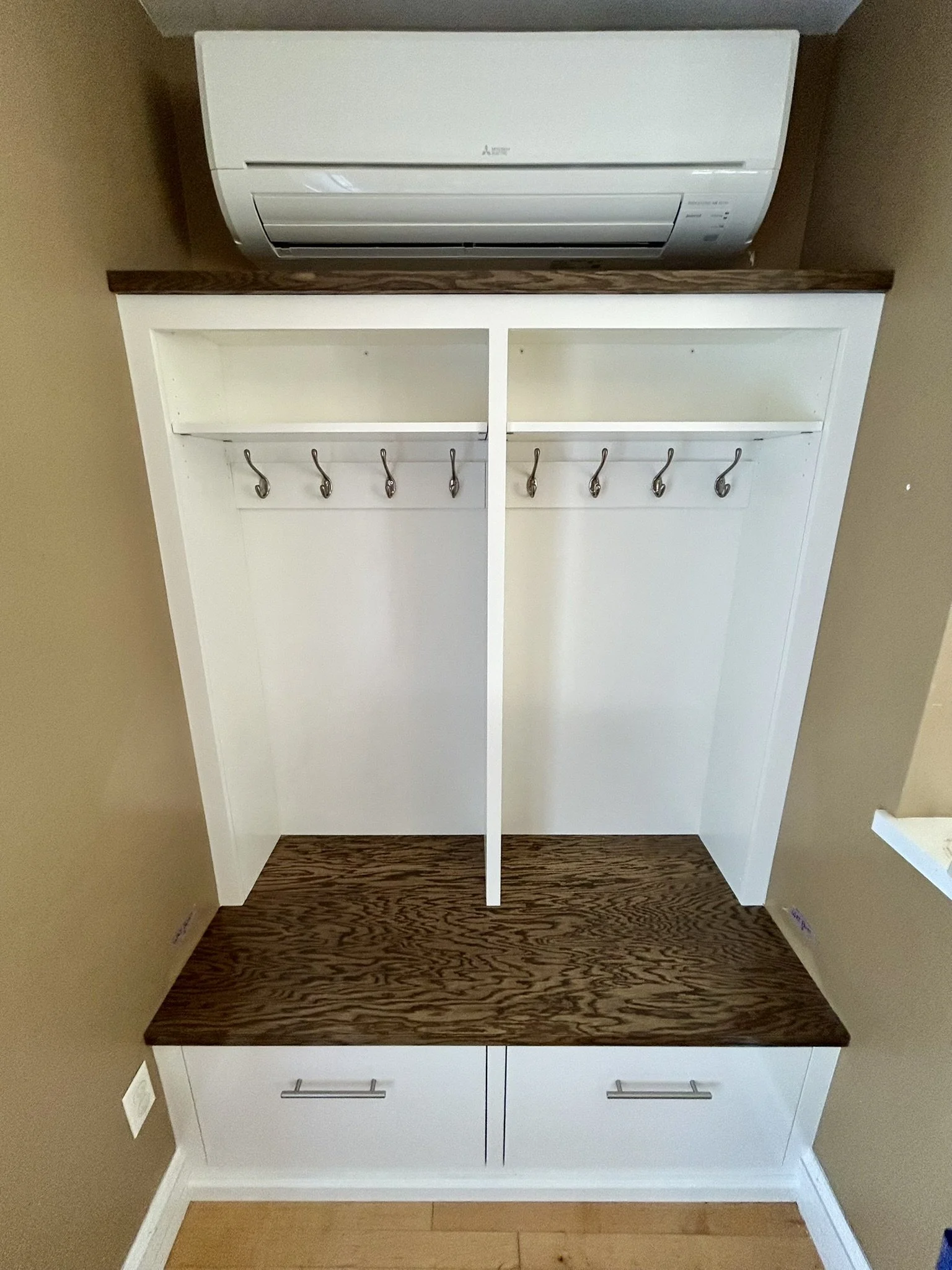 A white and dark brown entryway organizer with hooks for hanging items and a shelf with drawers below, mounted on a wall with an air conditioning unit above.