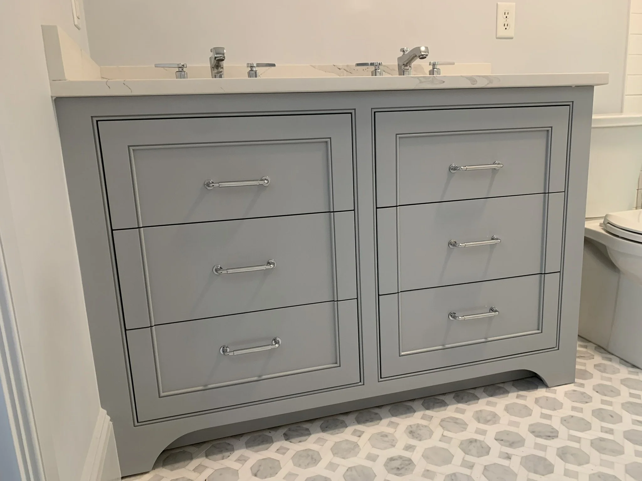 Gray bathroom vanity with six drawers, three on each side, topped with a white marble countertop and two faucets, in a bathroom with hexagonal patterned floor tiles.