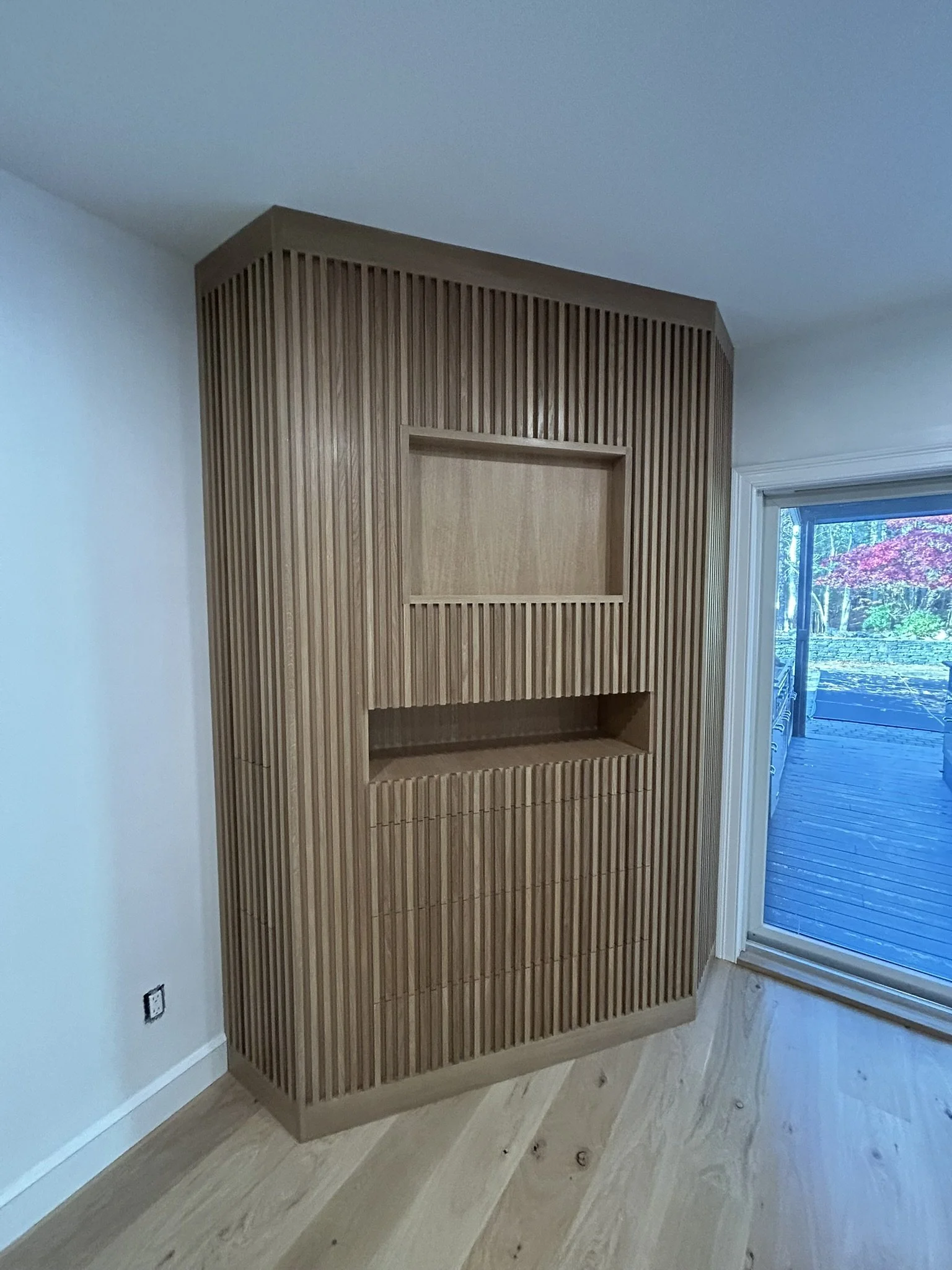 Interior of a room with a wooden accent wall featuring vertical slats and two recessed shelves, next to a sliding glass door with a view of an outdoor deck and garden.