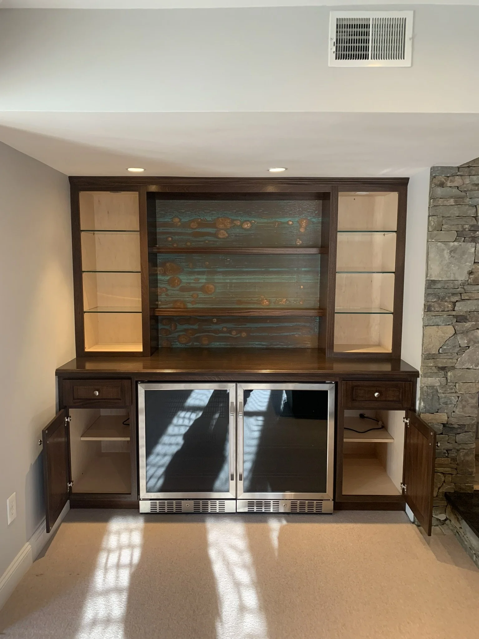 Empty built-in wooden cabinet with glass doors, open compartments, and a mini fridge, set against a wall with a stone accent on the right side.