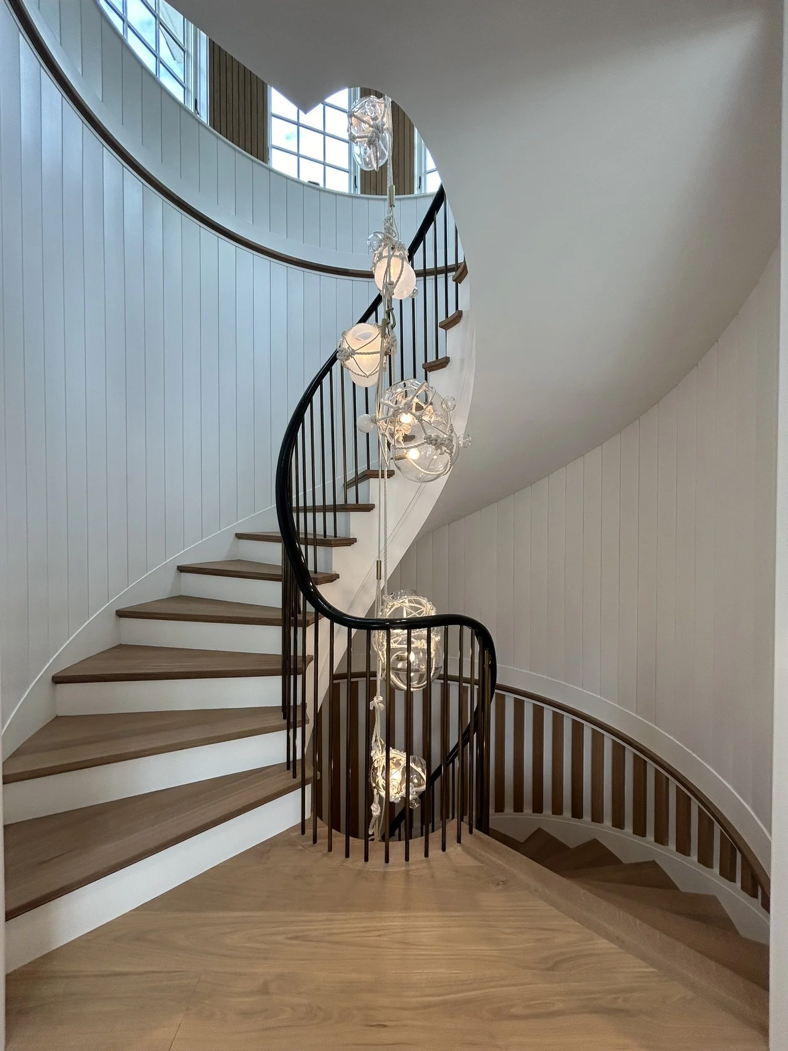 Interior view of a curved staircase with wooden steps and black railings, decorated with hanging glass and LED lights.