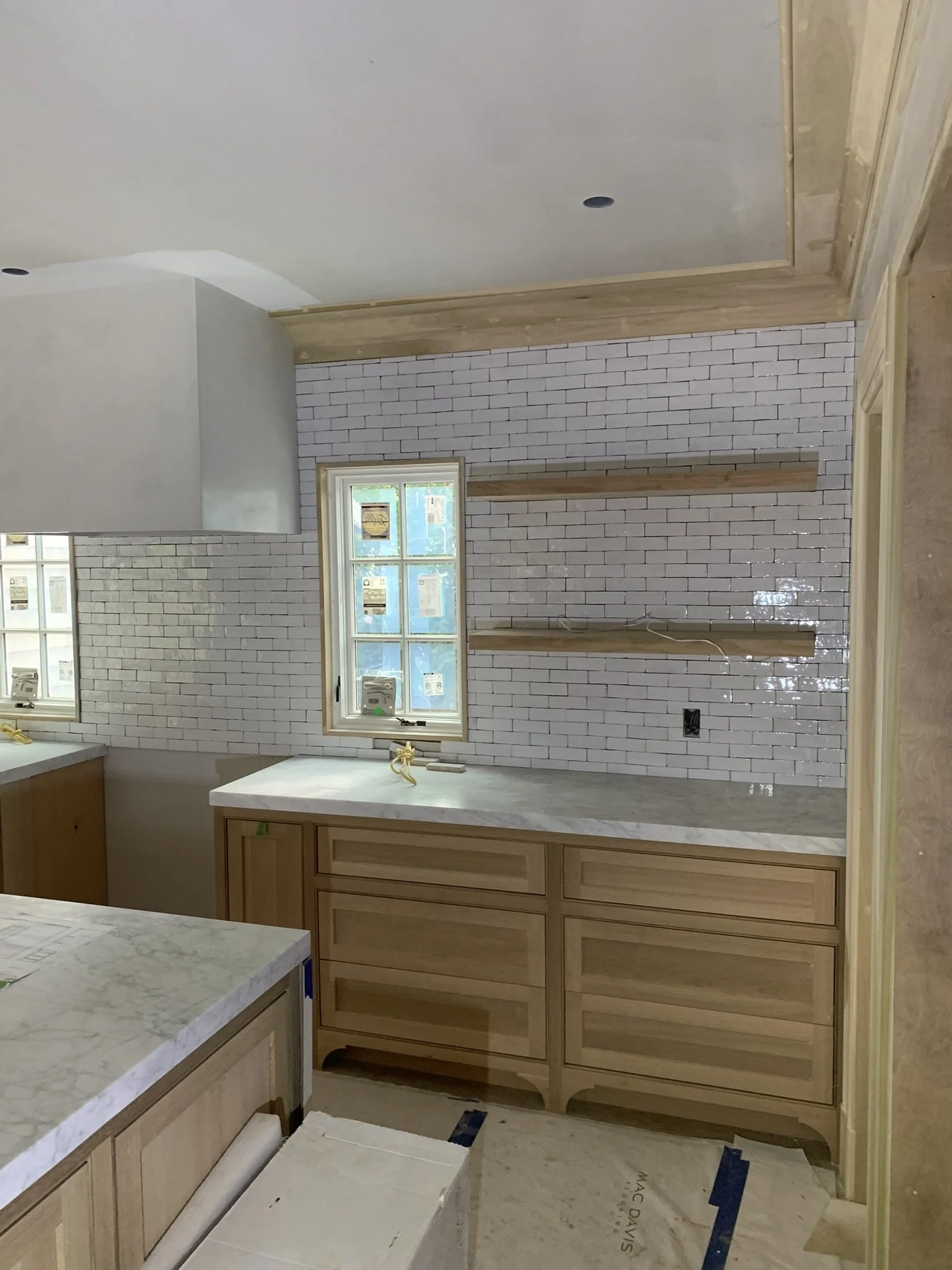 Kitchen under construction with white shiplap brick wall, wooden cabinets, floating wooden shelves, a window, and marble countertops.