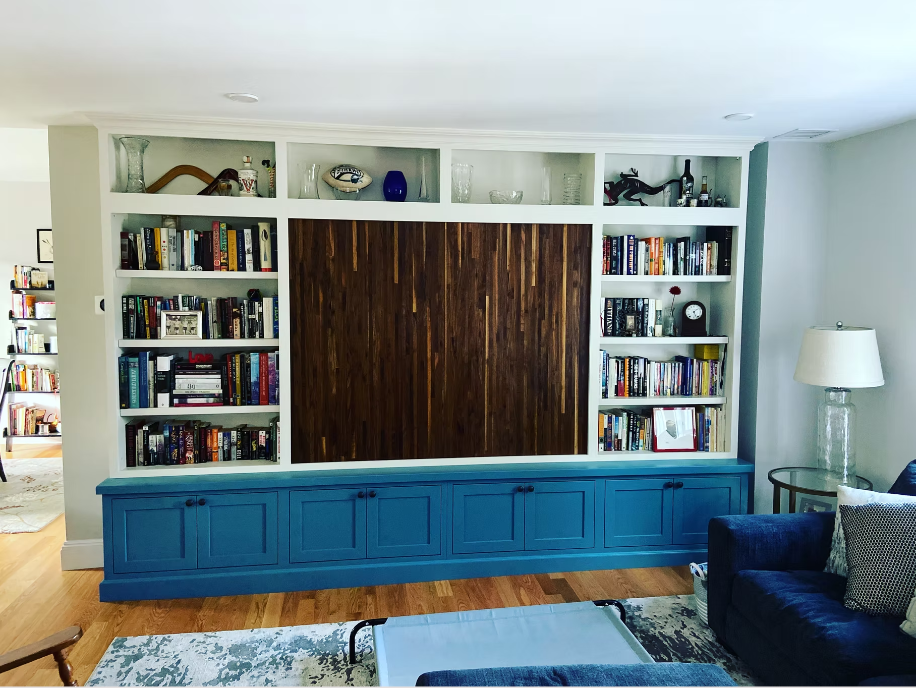 A living room with a built-in white bookshelf against the wall, containing books and decorative items. The lower part of the bookshelf is painted blue, with closed cabinets. There is a dark blue sofa with pillows to the right and a glass table in fro