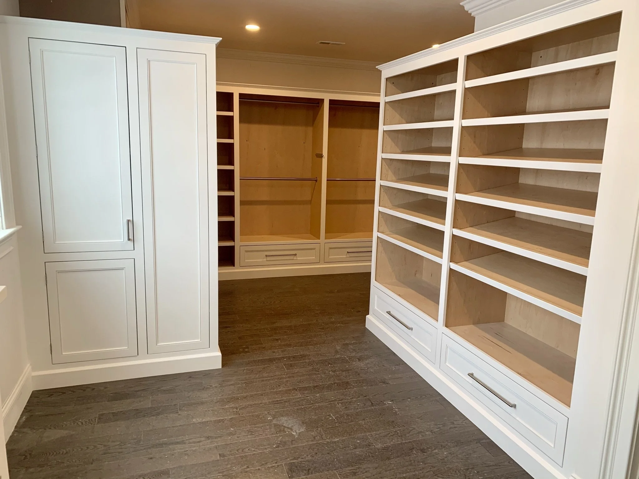 Empty walk-in closet with built-in white shelves and cabinets, with dark wooden flooring and overhead lighting.