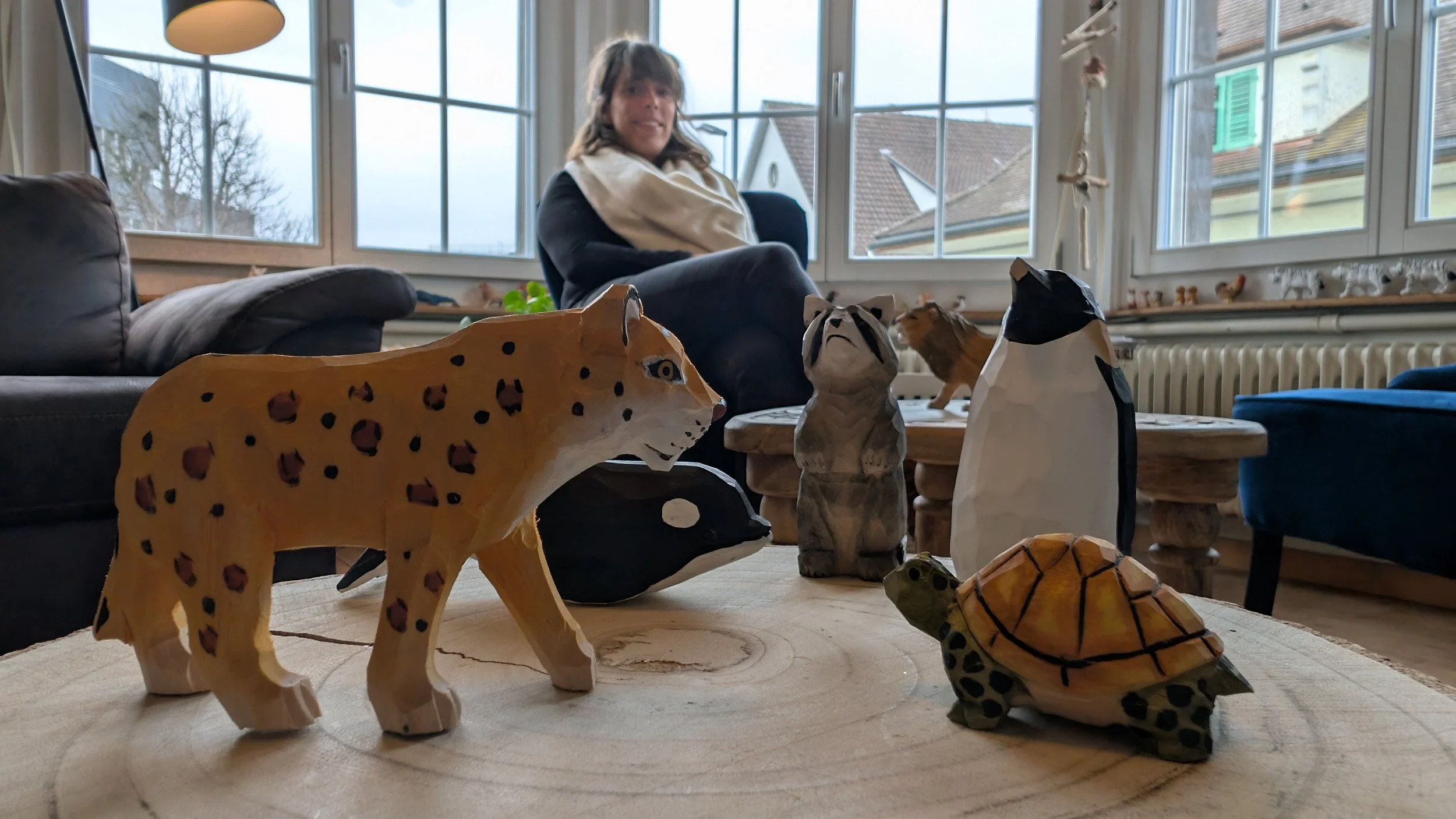 Holztiere auf einem Baumstamm, im Hintergrund eine Frau sitzt im Wohnzimmer mit großen Fenstern.