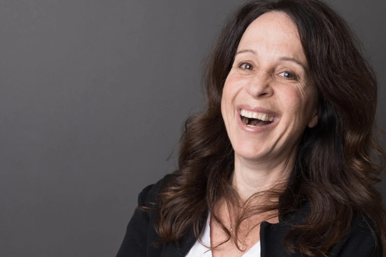 A woman with long, wavy dark brown hair smiling broadly against a gray background.