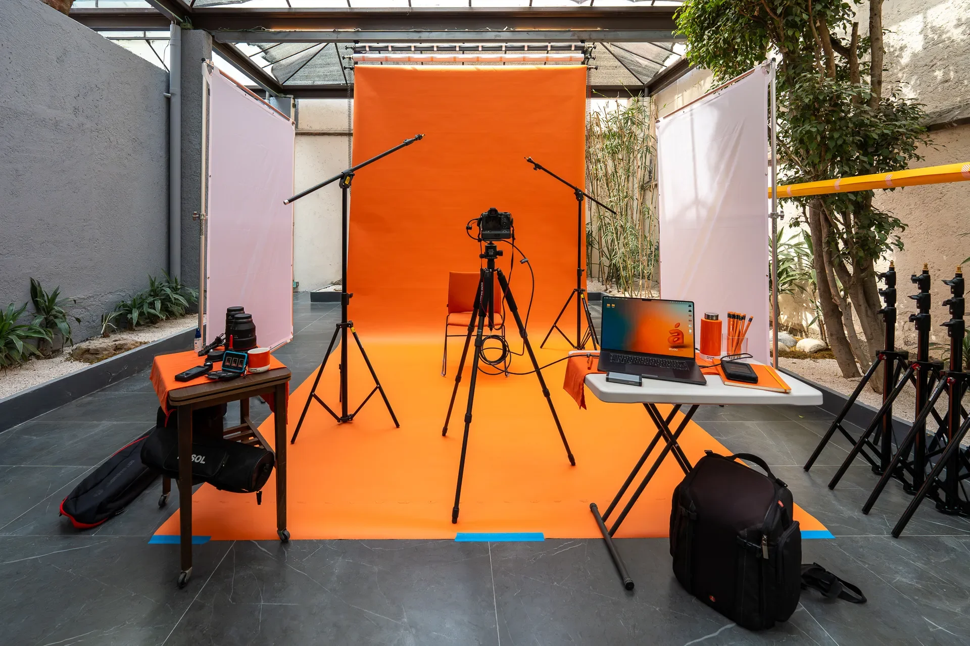 Estudio de grabación con fondo naranja, cámara, tripies, micrófonos y equipo de tecnología en un ambiente cubierto y decorado con plantas.