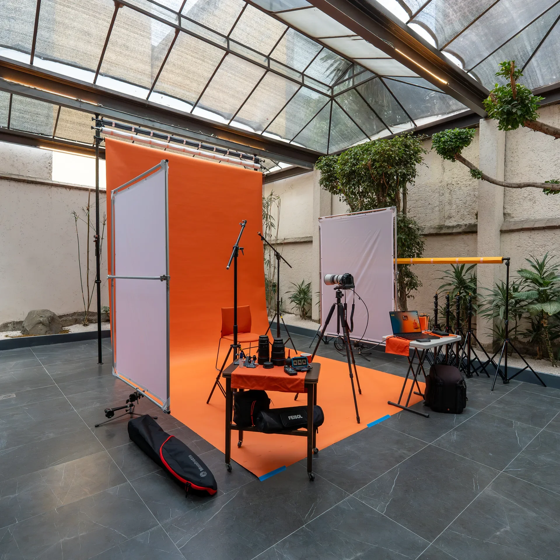 Estudio fotográfico con fondo naranja y blanco, equipo de cámara, trípodes, luces, y mesa con equipo en un espacio techado con plantas.