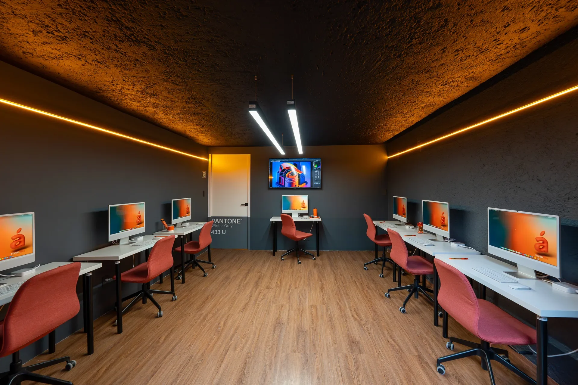 Aula de computadoras con mesas blancas, monitores y sillas rosadas, paredes grises y techo oscuro con iluminación decorativa, piso de madera.