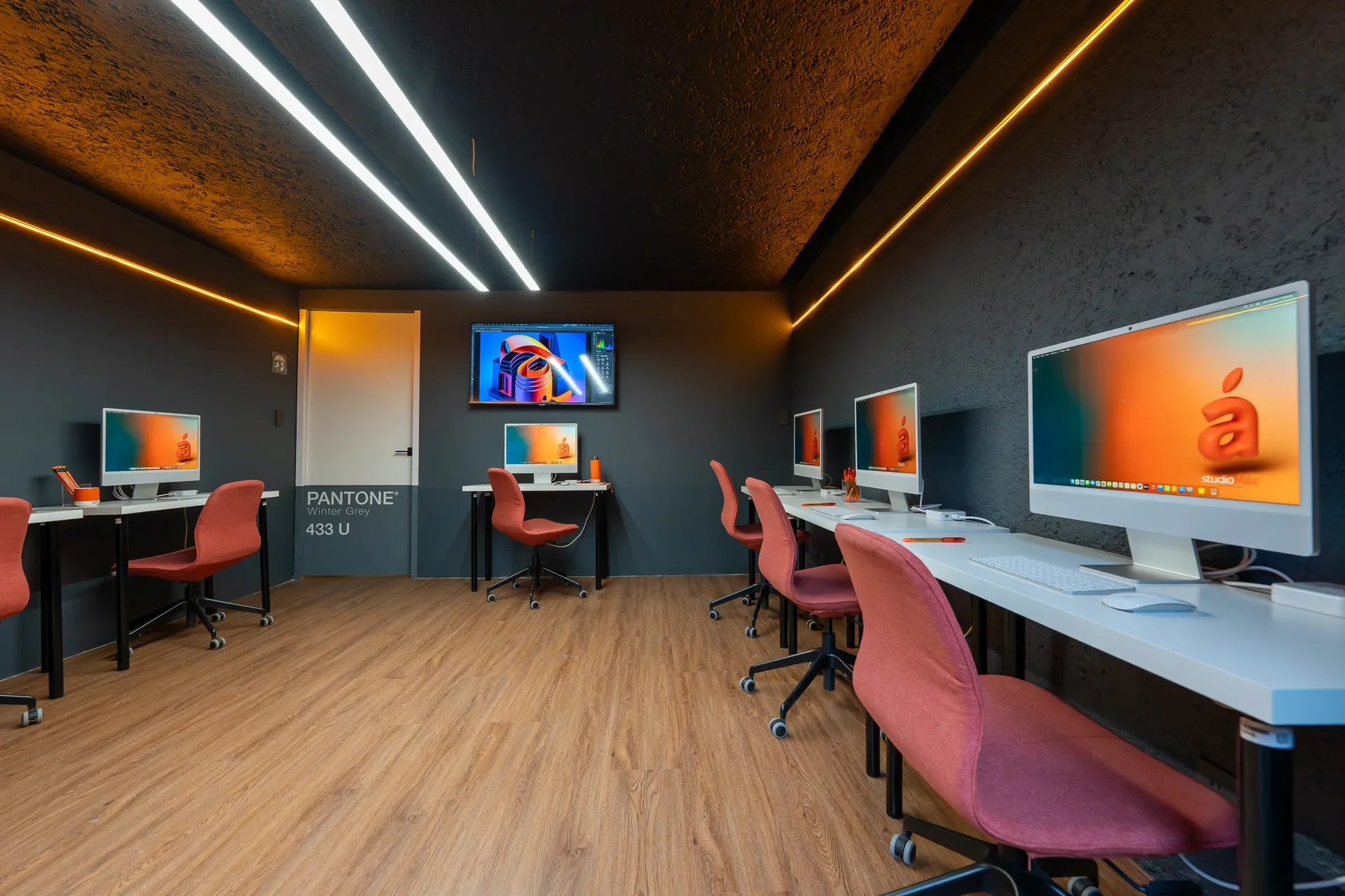 Sala de computadoras con varias estaciones de trabajo y pantallas modernas, sillas color durazno, paredes grises, piso de madera y luces LED en el techo.