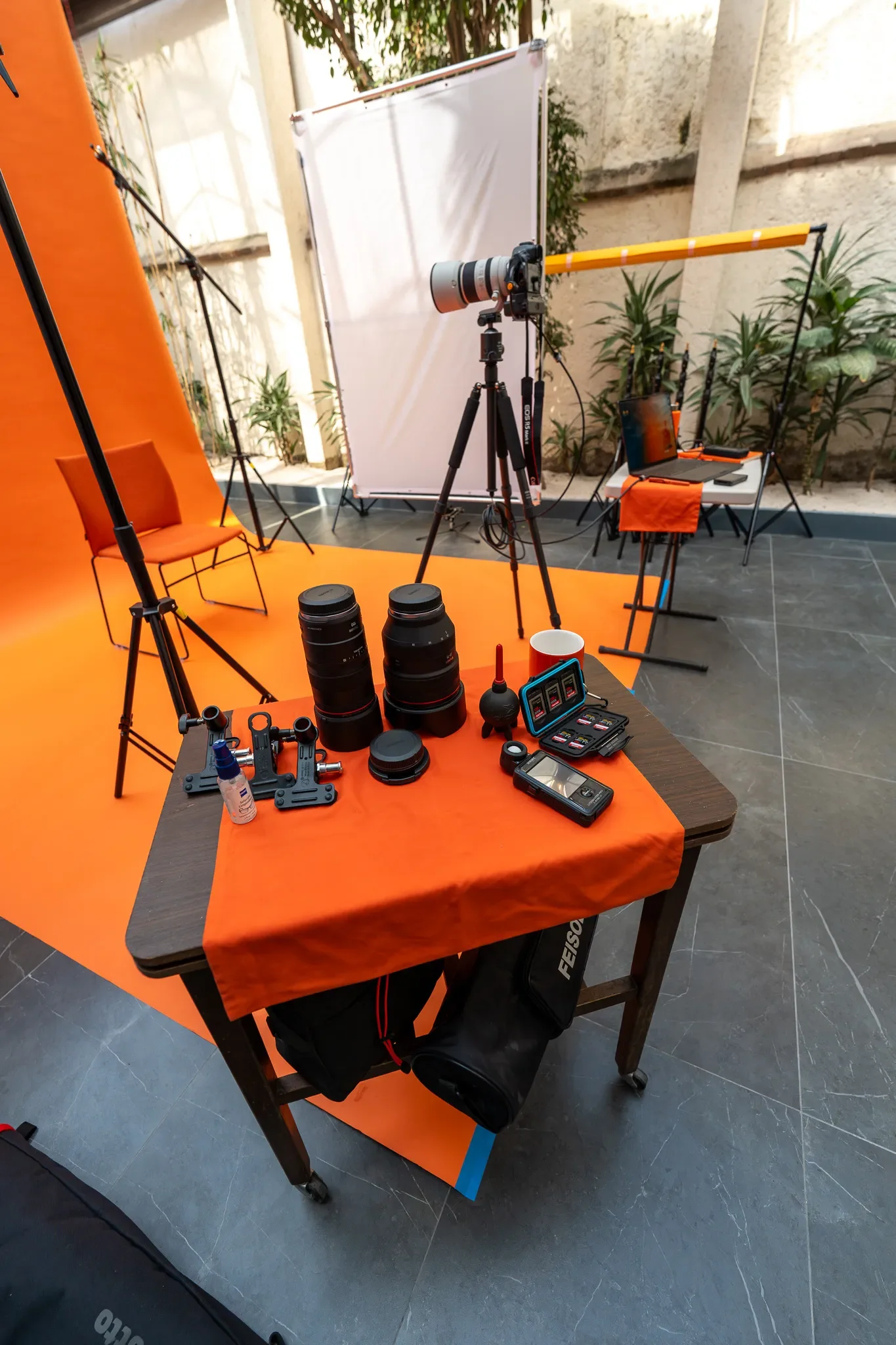 Equipo de fotografía y vídeo en un estudio con fondo anaranjado y cortina blanca, incluyendo cámaras, lentes, trípodes, luz de paraguas, y accesorios técnicos.