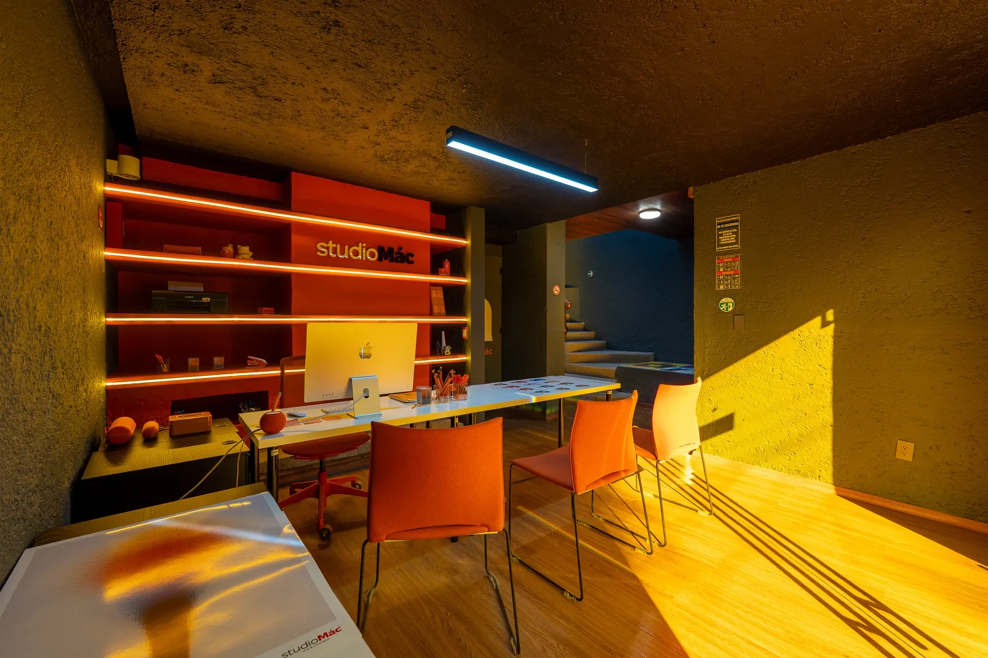 Oficina moderna con escritorio, computadora Apple, sillas naranjas, estantes con iluminación roja y señalización en la pared, con luz natural entrando por la ventana.