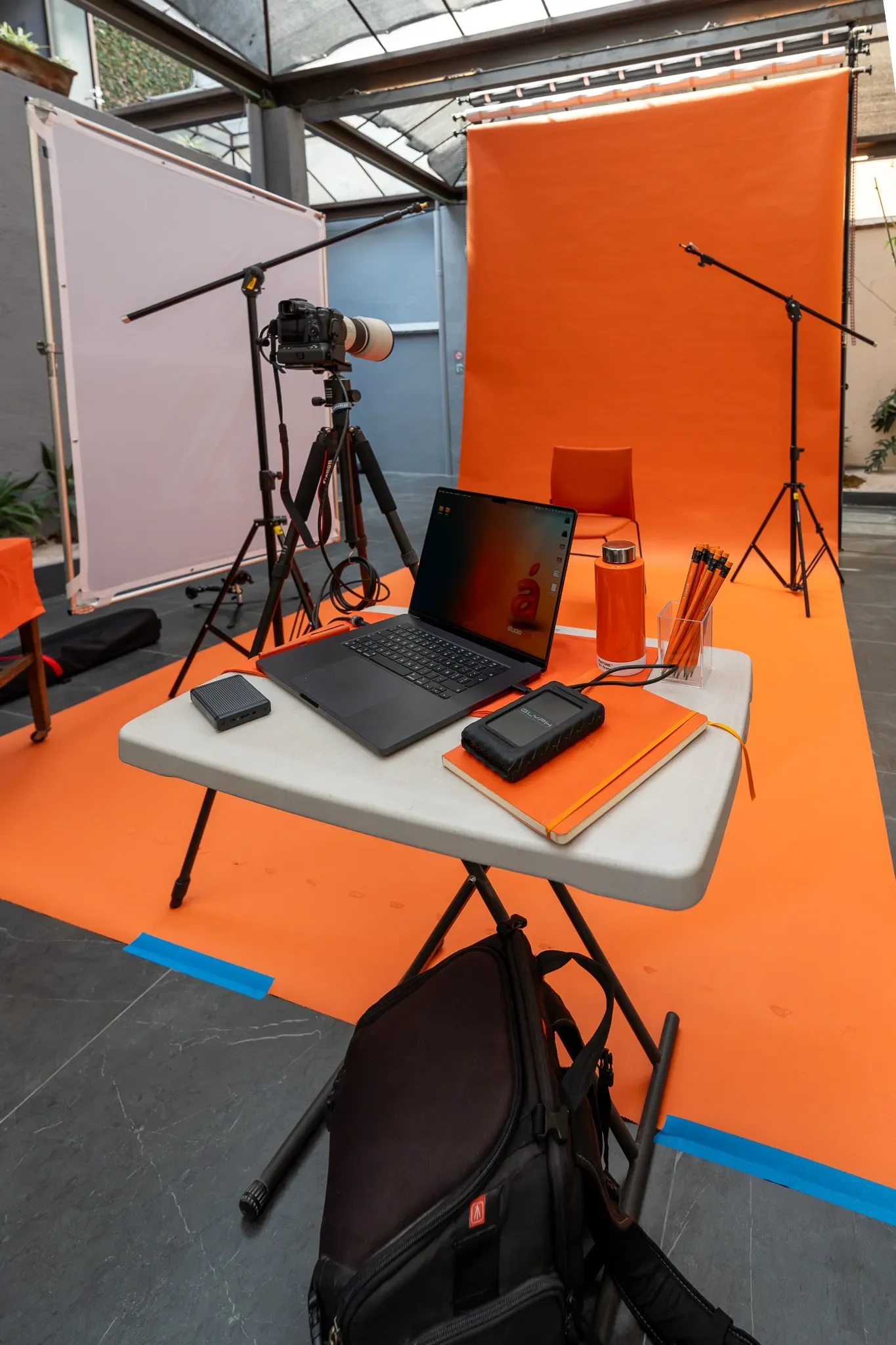 Un set de grabación de foto de estudio con fondo naranja, equipo de cámaras, luz, laptop, cuadernos, termos y otros accesorios en un espacio interior.
