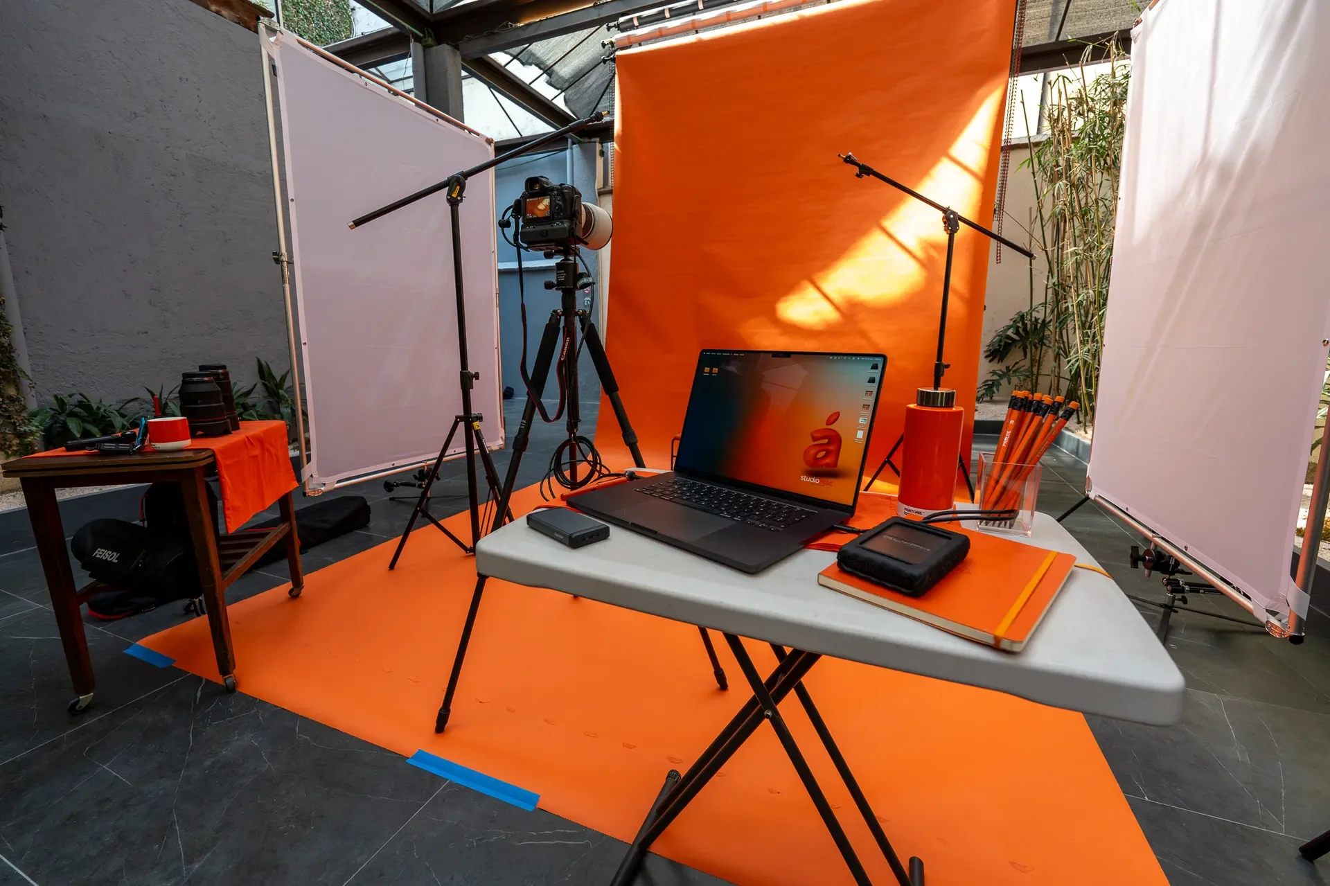 Estudio de grabación con fondo naranja, cámara en trípode, luces, laptop, y materiales de oficina en mesa y mesa auxiliar, en un espacio con plantas y estructura de vidrio