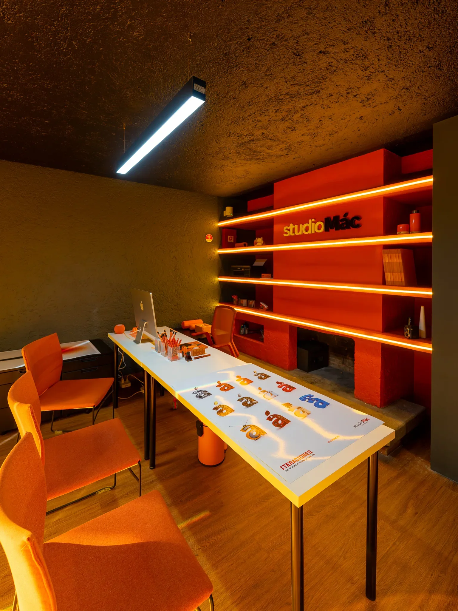 Oficina moderna con una mesa blanca, sillas naranjas, estanterías con iluminación de neón naranja y un cartel que dice 'studio Mác' en la pared roja.